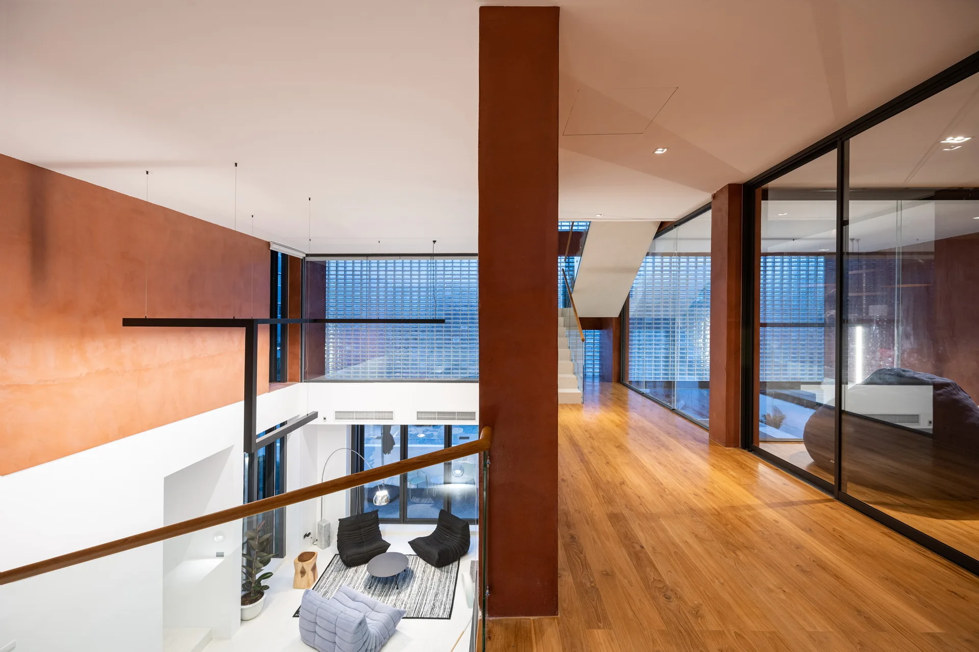 An upper mezzanine landing with warm wood floors and terracotta-painted columns overlooks a lower-level living room with a glass block wall.