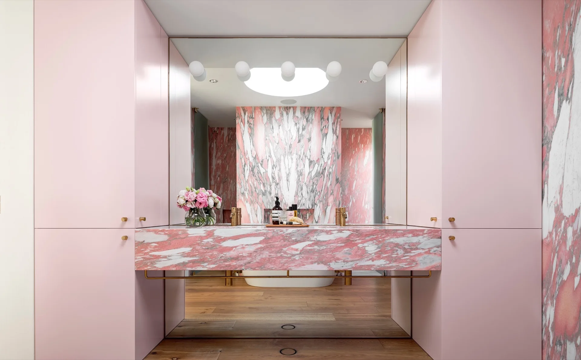 A bathroom vanity features a pink marble countertop flanked by tall pink cabinets, with a large mirror reflecting a matching marble wall and bathtub.