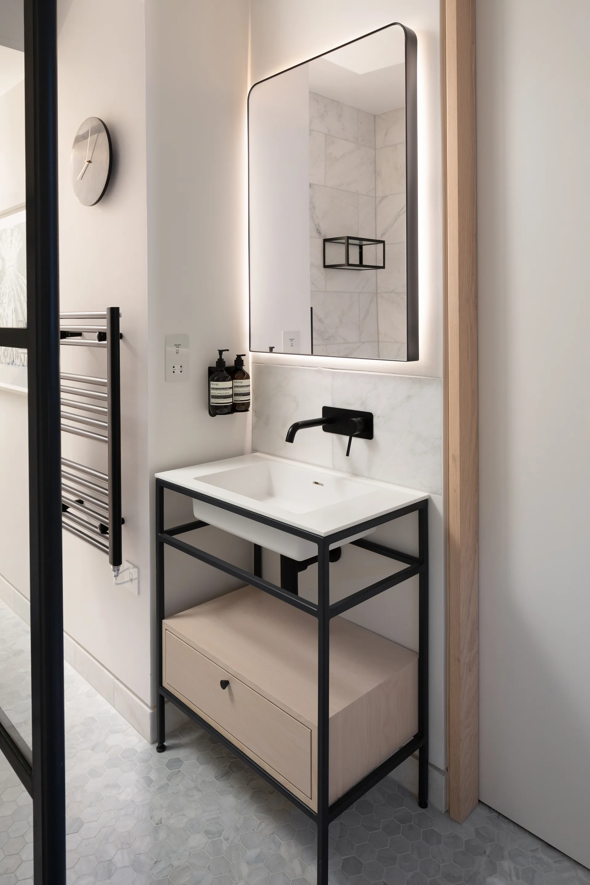 A modern bathroom features a black metal vanity frame with a wood drawer, a backlit rectangular mirror, and marble hexagon floor tiles.