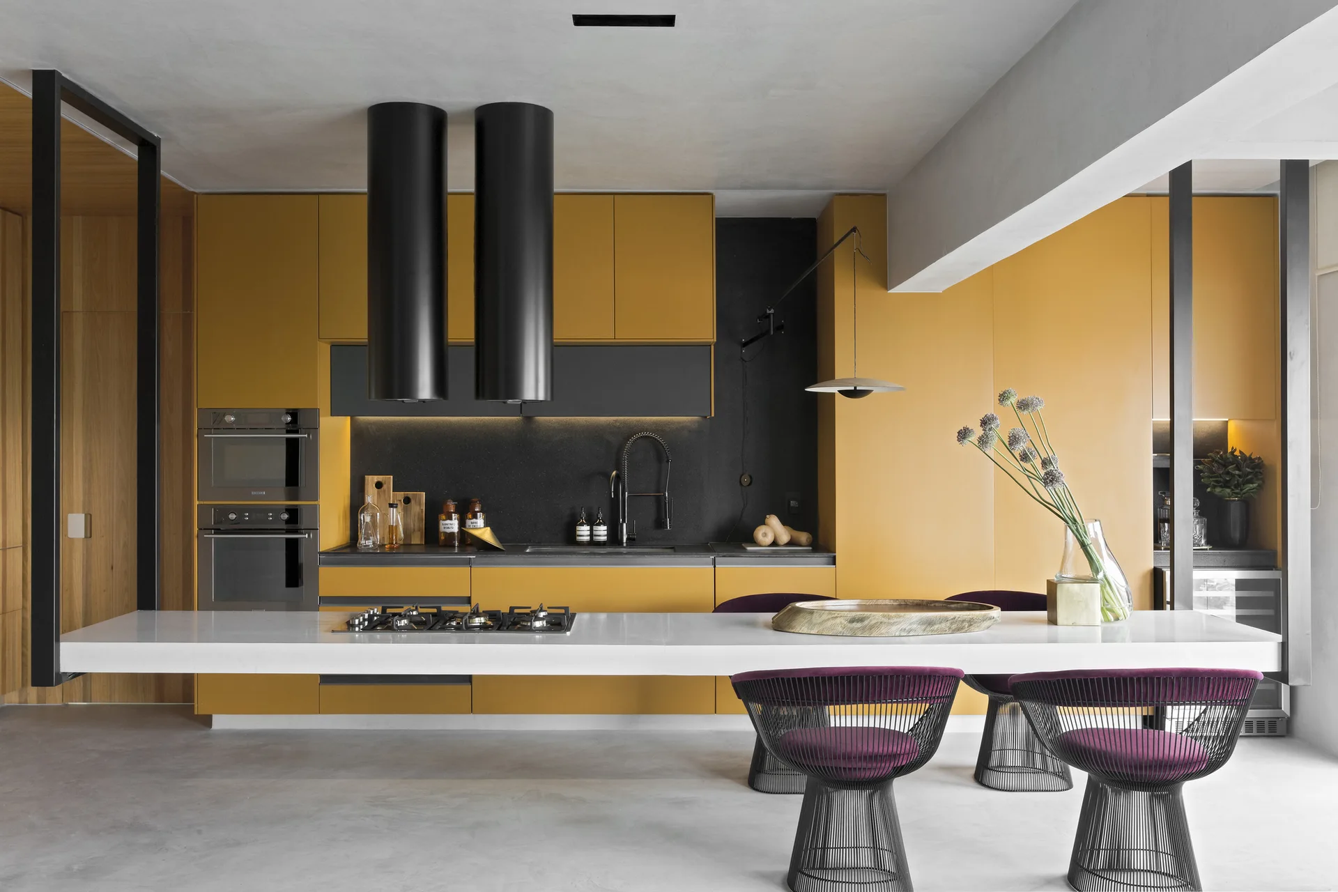 A modern kitchen featuring matte mustard yellow cabinetry, two black cylindrical range hoods, and a long white island with purple wire-frame stools.