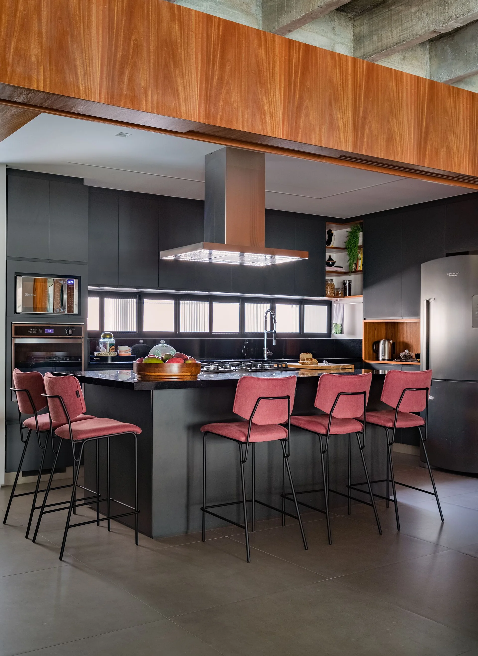 A modern kitchen features matte black cabinets, a central island with pink upholstered stools, and a warm wood soffit under concrete ceiling beams.