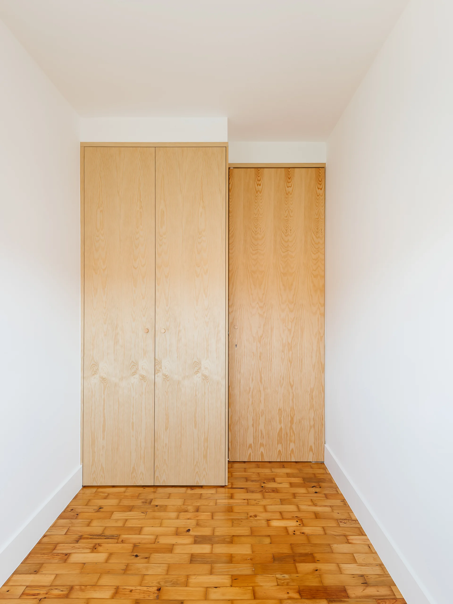 Minimalist built-in closet doors with natural wood grain are set flush into white walls above detailed wood parquet flooring.