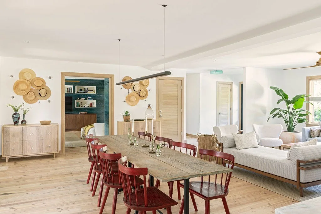 An open-concept dining space features a long wooden table with red chairs, a neutral chaise lounge, and straw hats hung on the white wall.