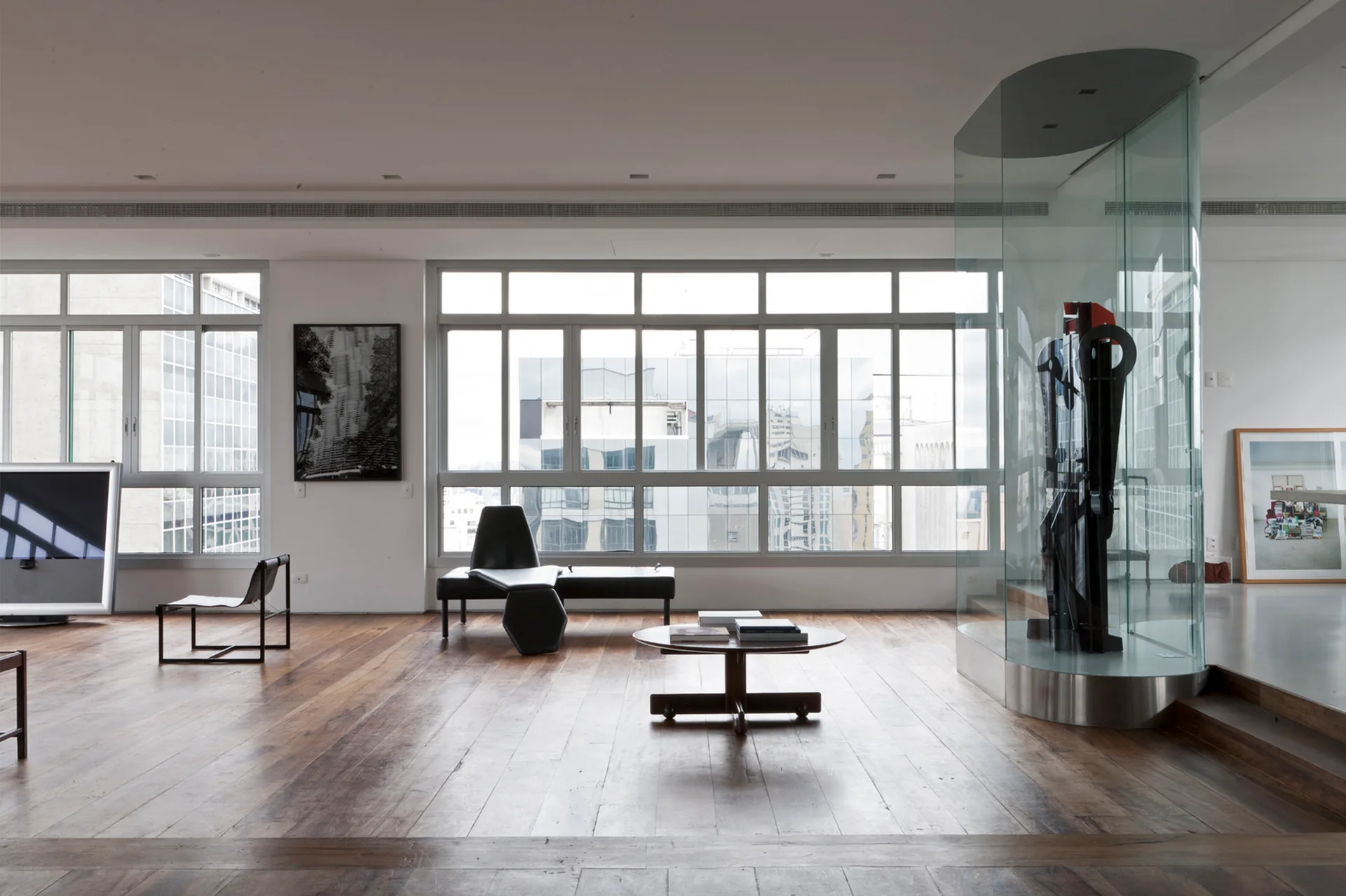 A spacious minimalist living room with rustic wood flooring, a low black lounge bench, and a tall curved glass art display case.
