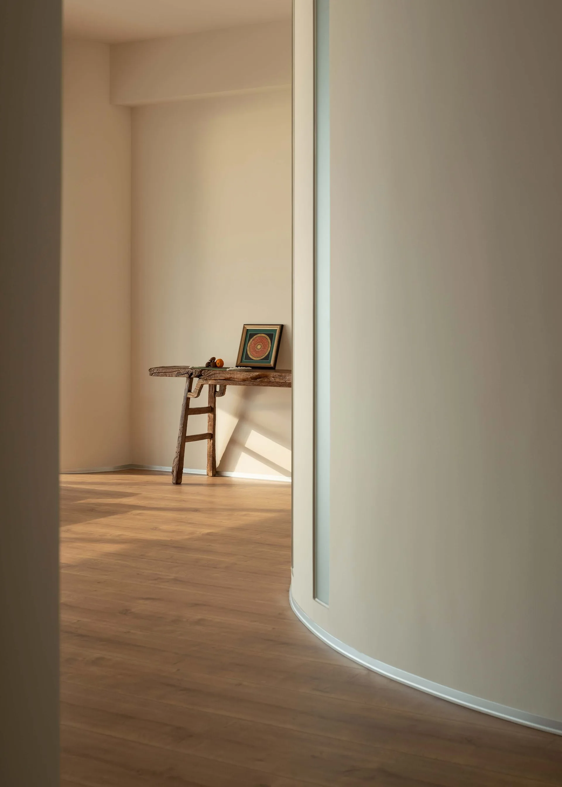 A minimalist beige hallway features a curved plaster wall and a rustic weathered wood console table displaying a small framed artwork.