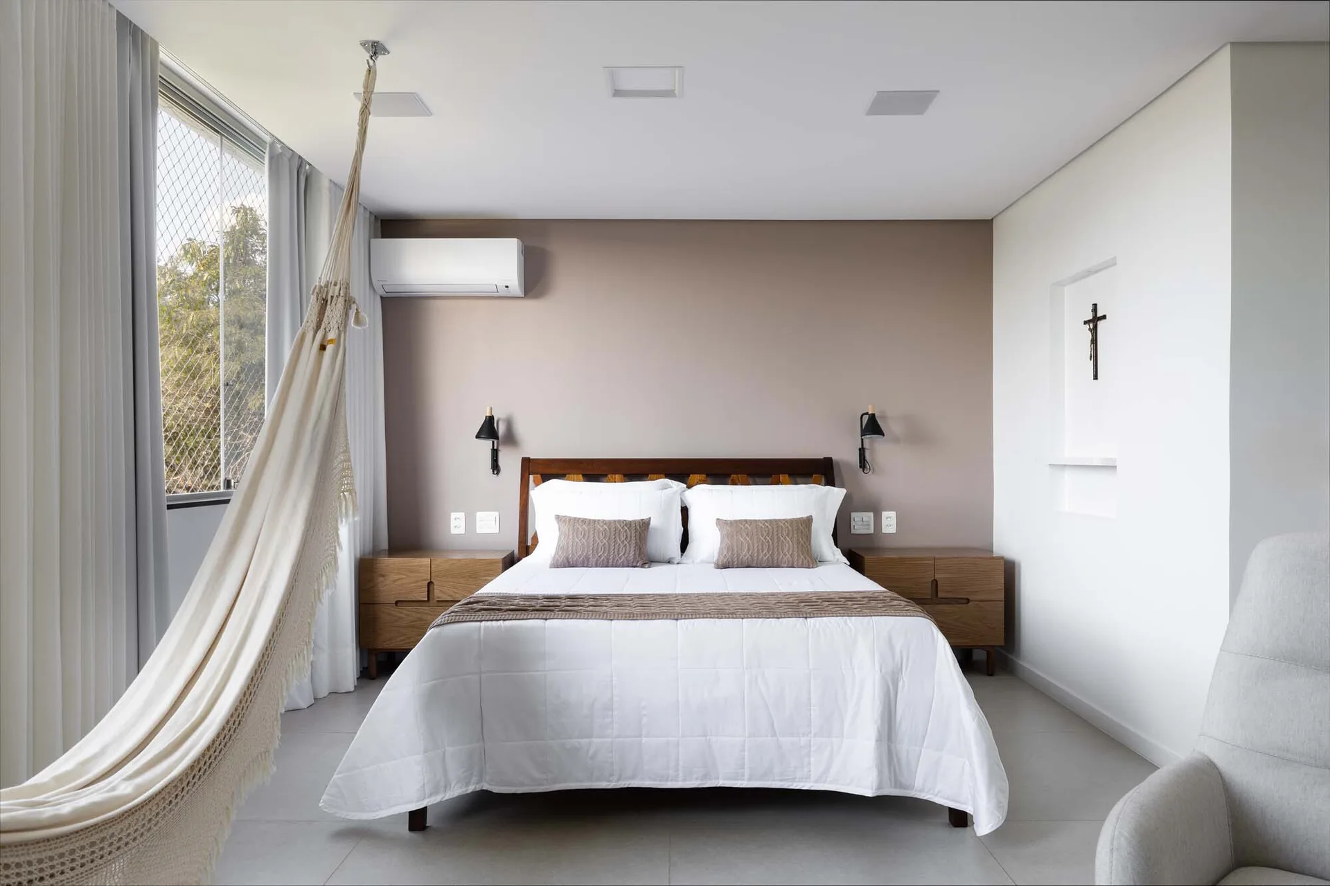 A modern bedroom features a bed flanked by wooden nightstands against a taupe accent wall, with a cream hammock hanging near a window on the left.