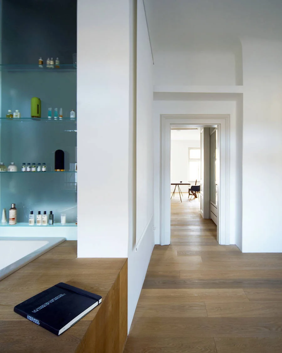 A minimalist bathroom entry featuring a recessed blue glass shelving niche, a light wood ledge, and an open doorway leading to a bright room.