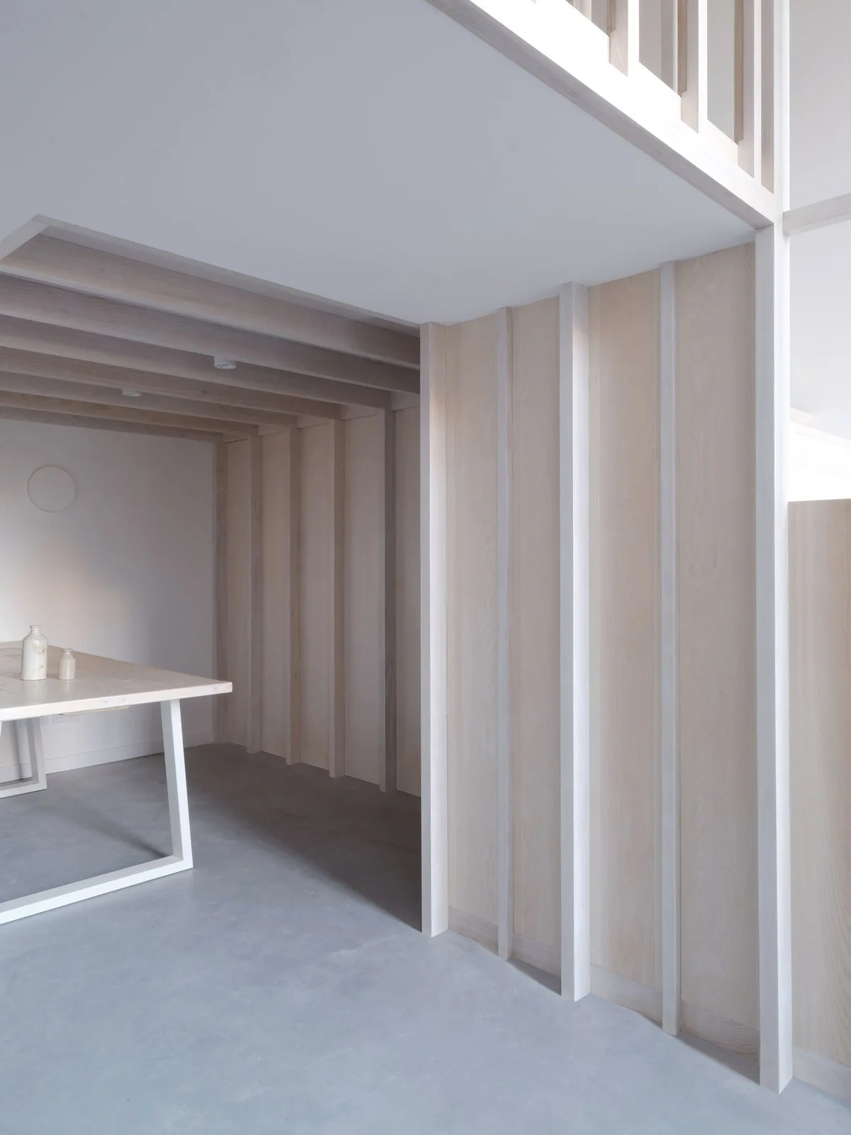 A minimalist room features light wood vertical wall paneling, matching ceiling beams, a grey concrete floor, and a wooden table with a white frame.
