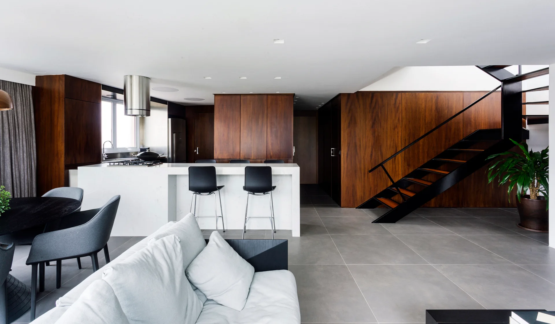 An open-concept living area with a white stone kitchen island, dark wood wall paneling, a grey tiled floor, and a black metal staircase.