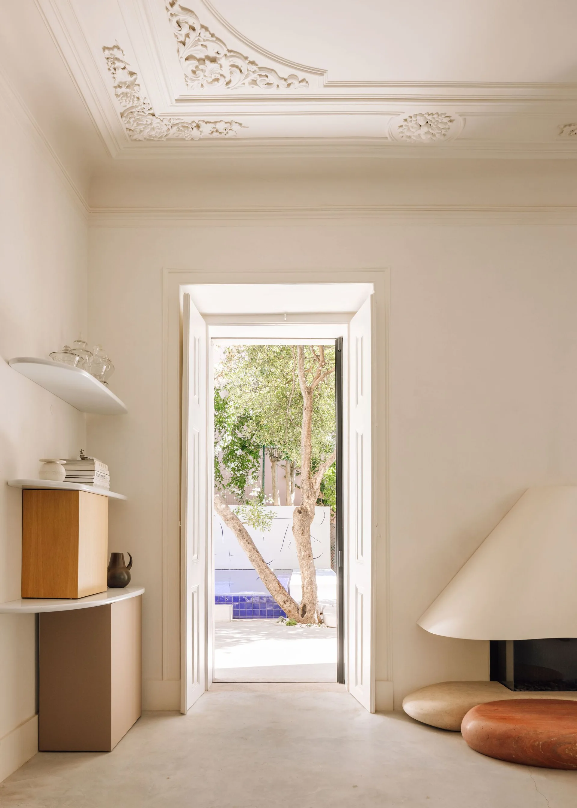 An off-white room features an ornate plaster ceiling, a minimalist sculptural fireplace with stone hearth seats, and open doors leading to a courtyard.