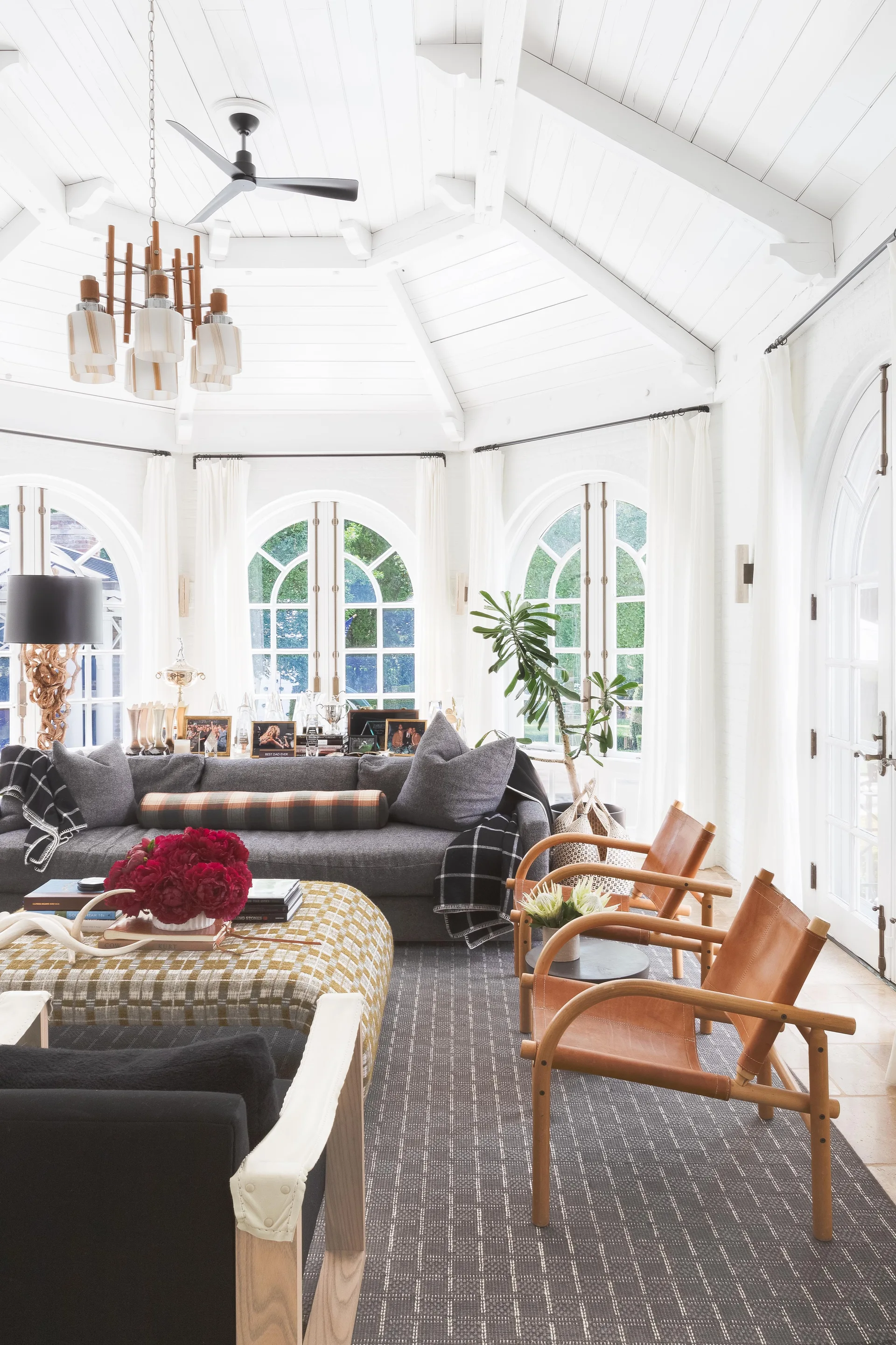 A sunlit living room features a grey sofa, tan leather accent chairs, a plaid ottoman, and large arched windows under a white vaulted ceiling.