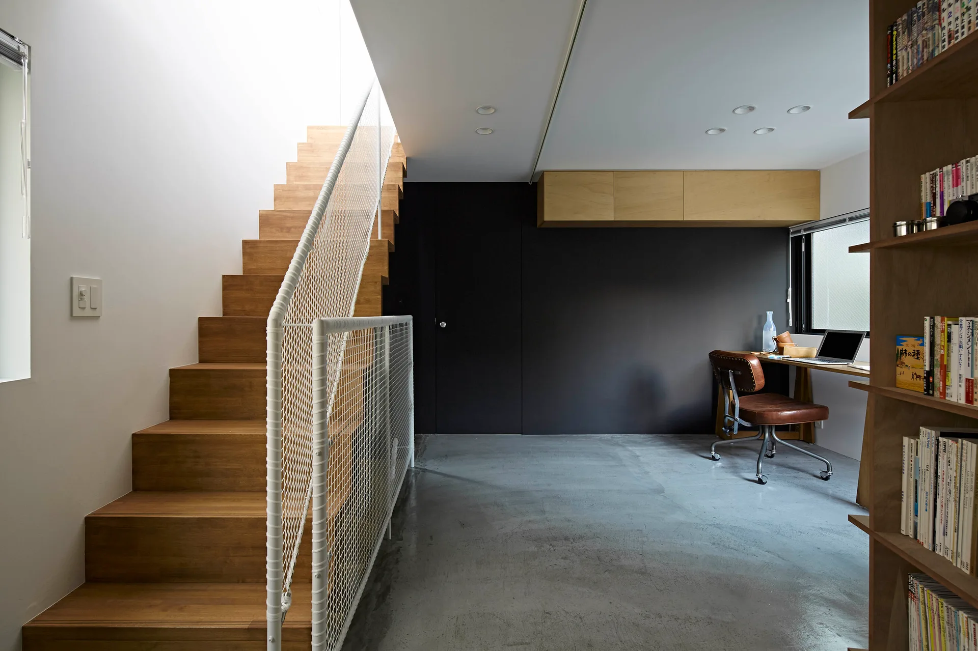 A minimalist home office features a wooden staircase with a white mesh railing, a concrete floor, and a built-in wooden desk against a dark wall.