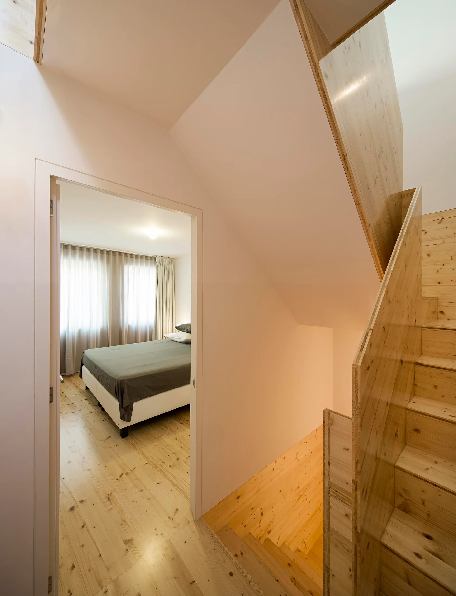 A light wood staircase with solid wood railings next to an open doorway showing a bedroom with grey bedding.