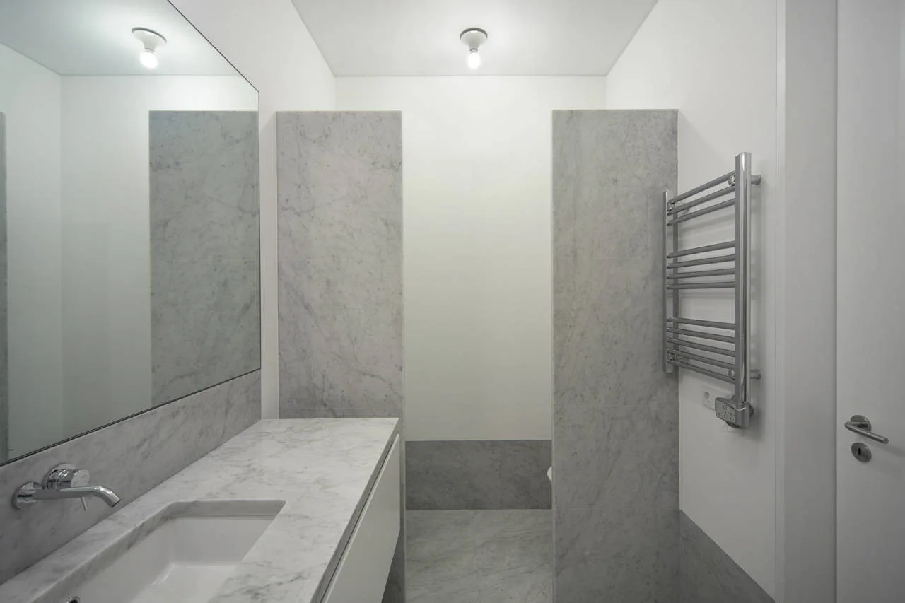 A minimalist bathroom featuring a gray marble vanity countertop, matching marble wall panels, a chrome heated towel rack, and white walls.