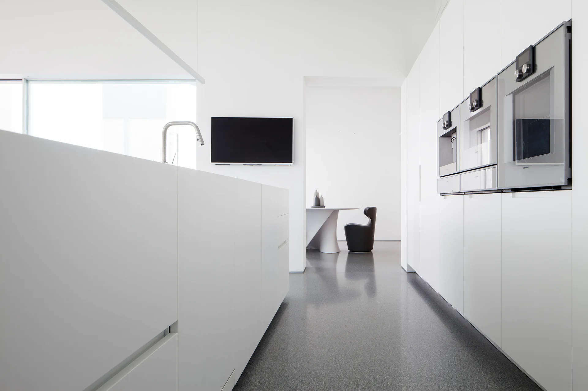 A minimalist white kitchen features seamless, handleless cabinetry, integrated silver wall ovens, and a highly reflective polished grey floor.