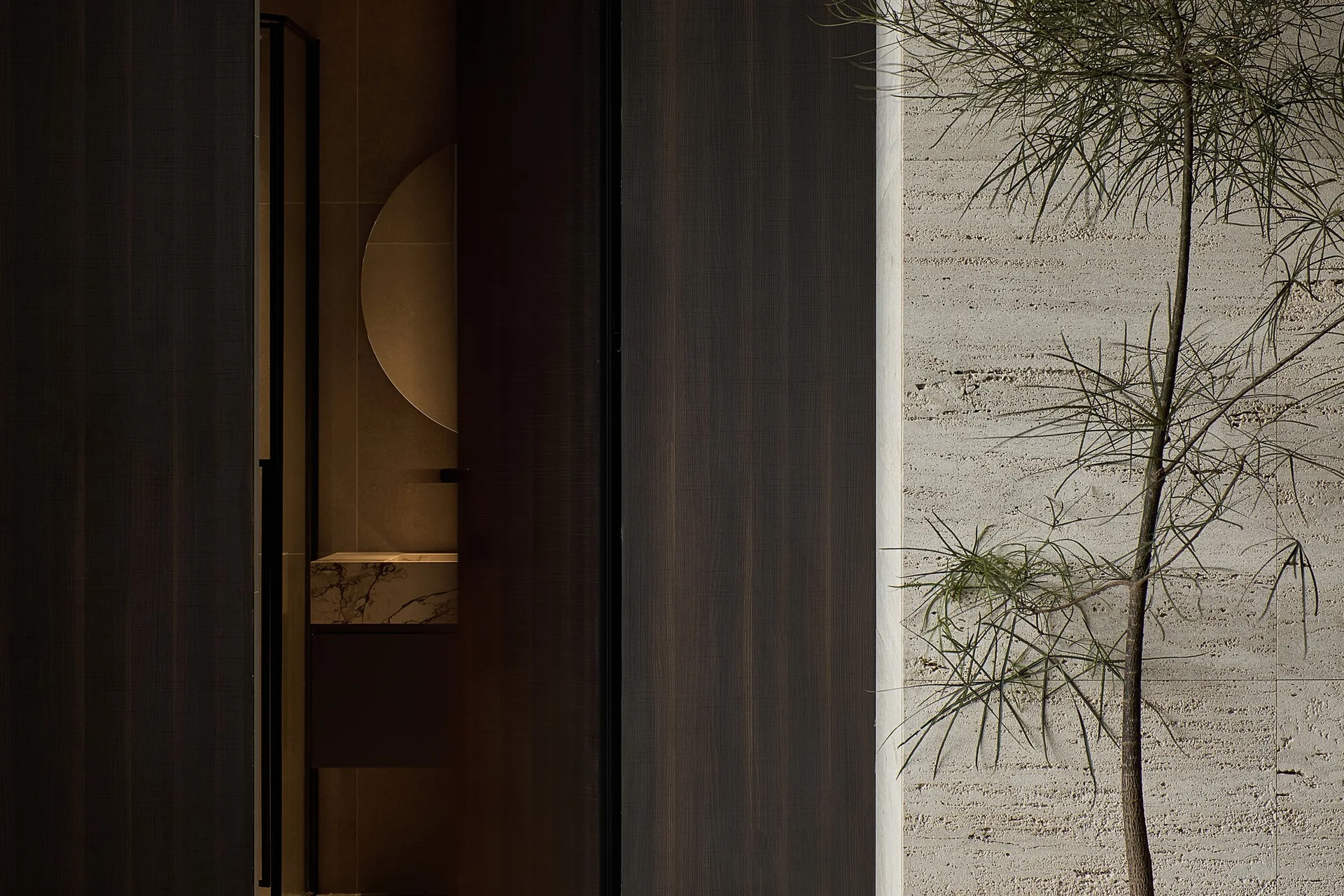 A dark wood sliding door reveals a glimpse of a modern bathroom with a marble vanity, circular mirror, and textured beige stone wall.