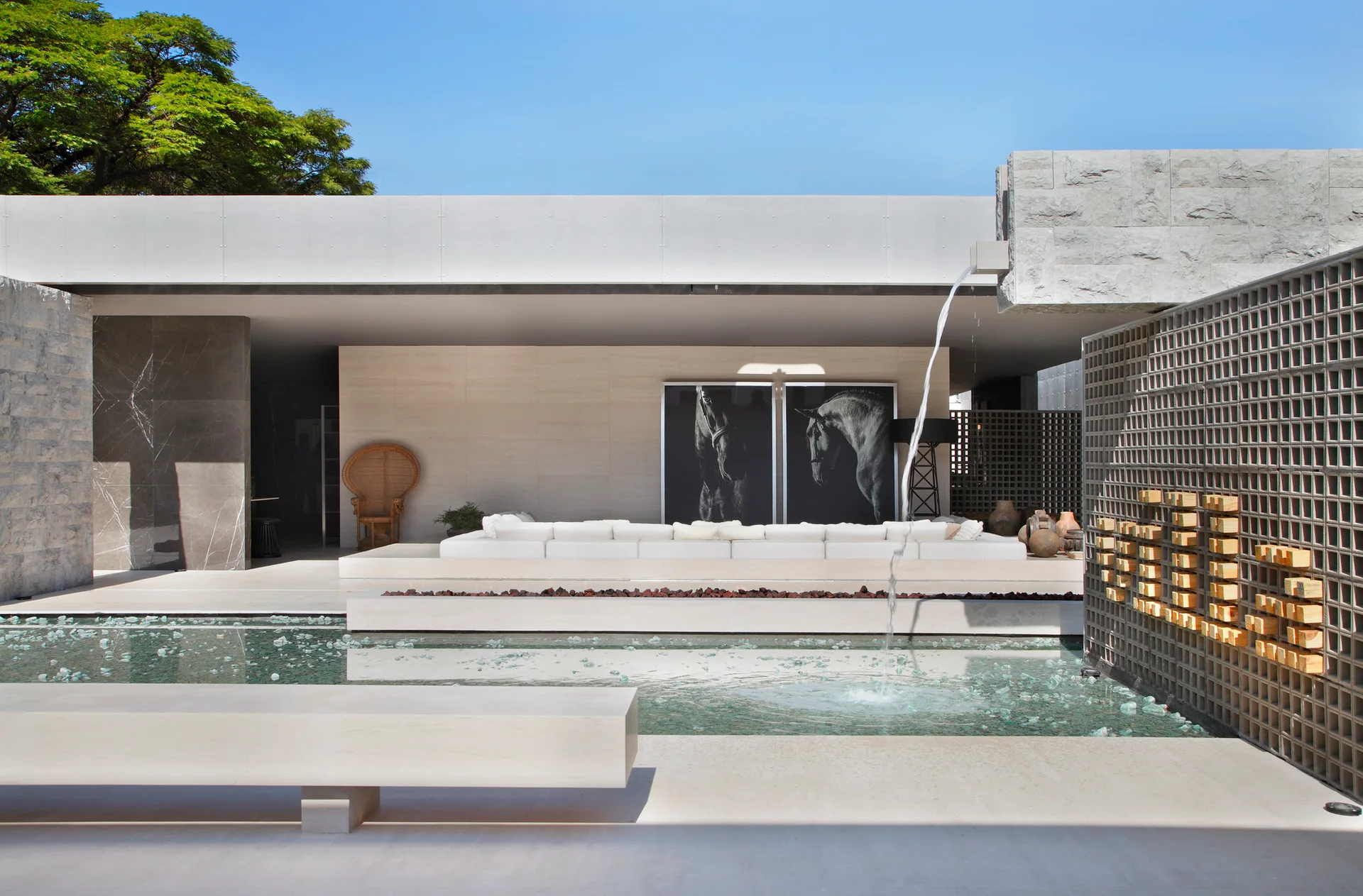 A modern outdoor patio featuring a white sectional sofa, a water fountain cascading into a pool, and a gray concrete breeze block wall.