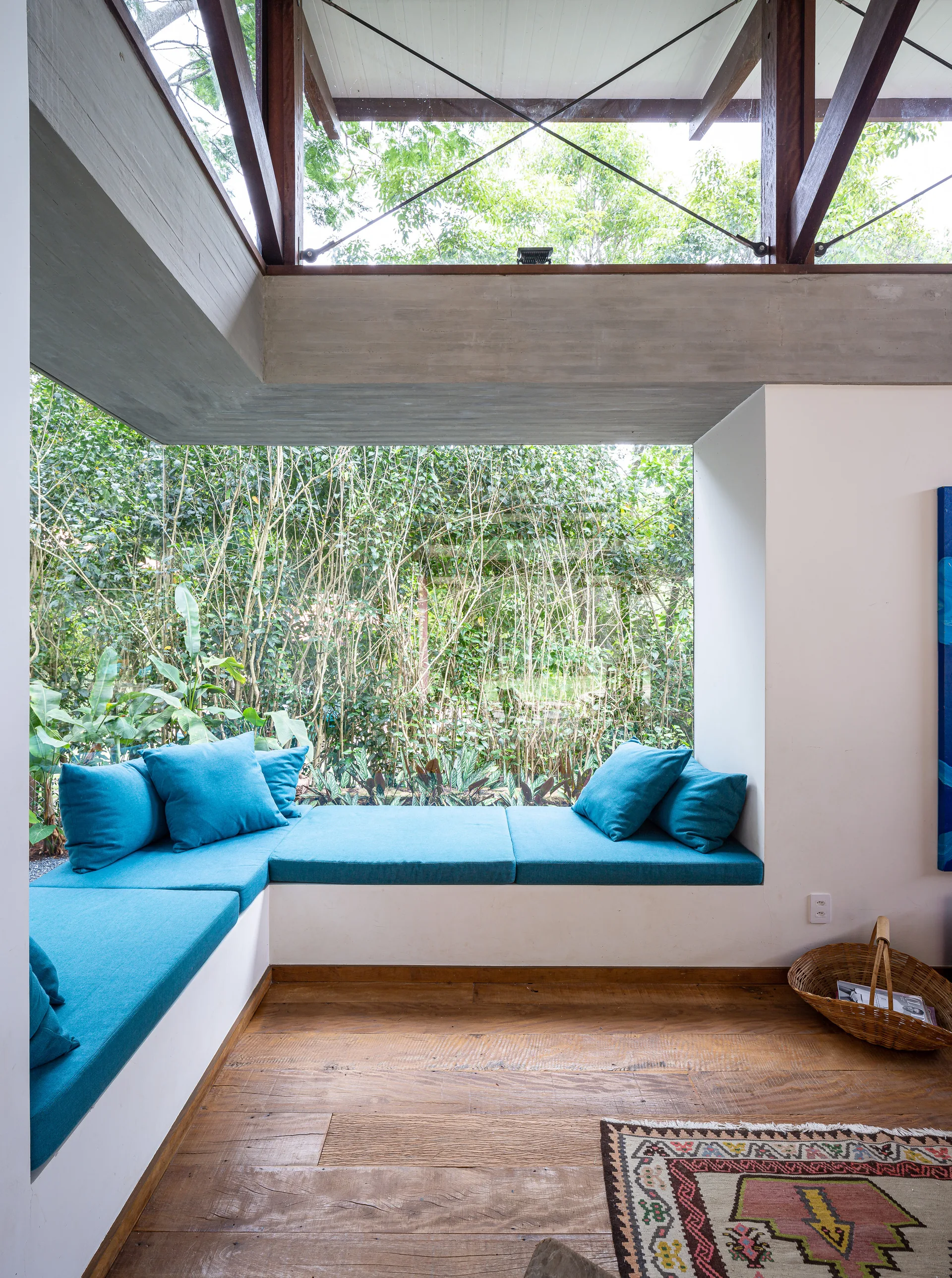 A built-in corner bench with teal cushions sits below a large window looking out at a lush garden in a room with wood flooring.