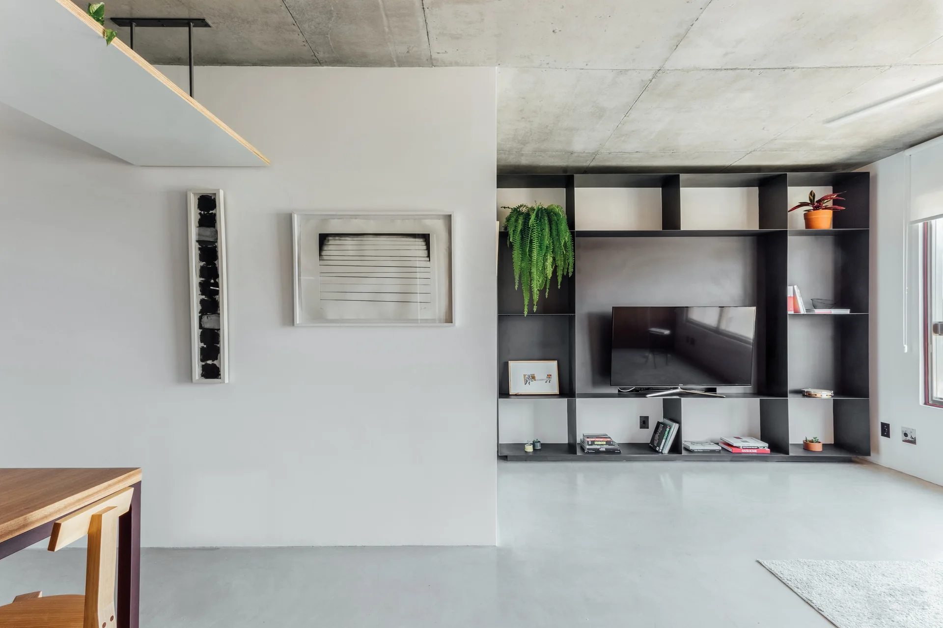 A minimalist living room features an exposed concrete ceiling, a large black metal grid shelving unit with a TV, and framed art on a white wall.