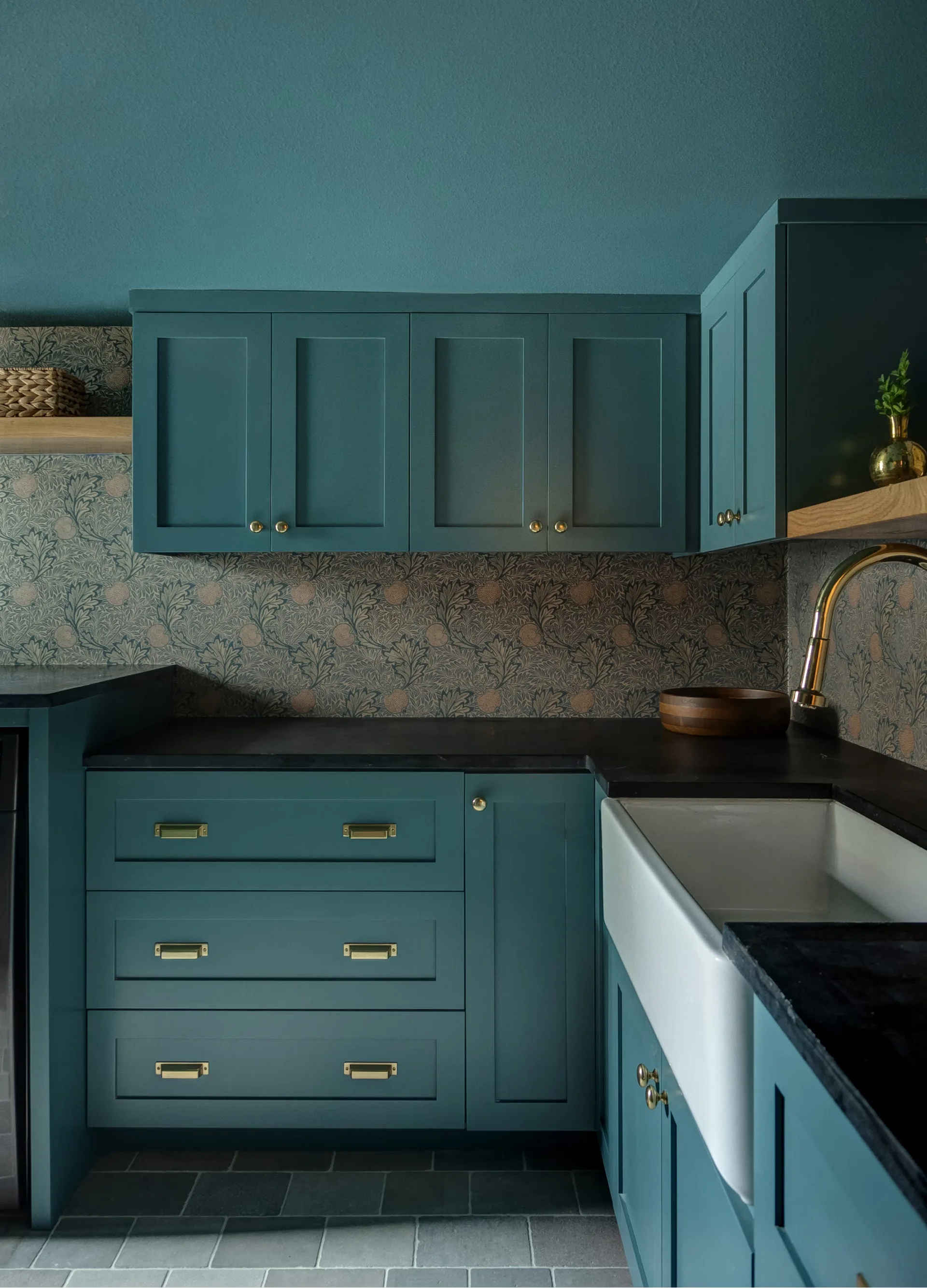A utility room corner featuring dark teal cabinetry, a botanical floral wallpaper backsplash, a white farmhouse sink, and a black countertop.