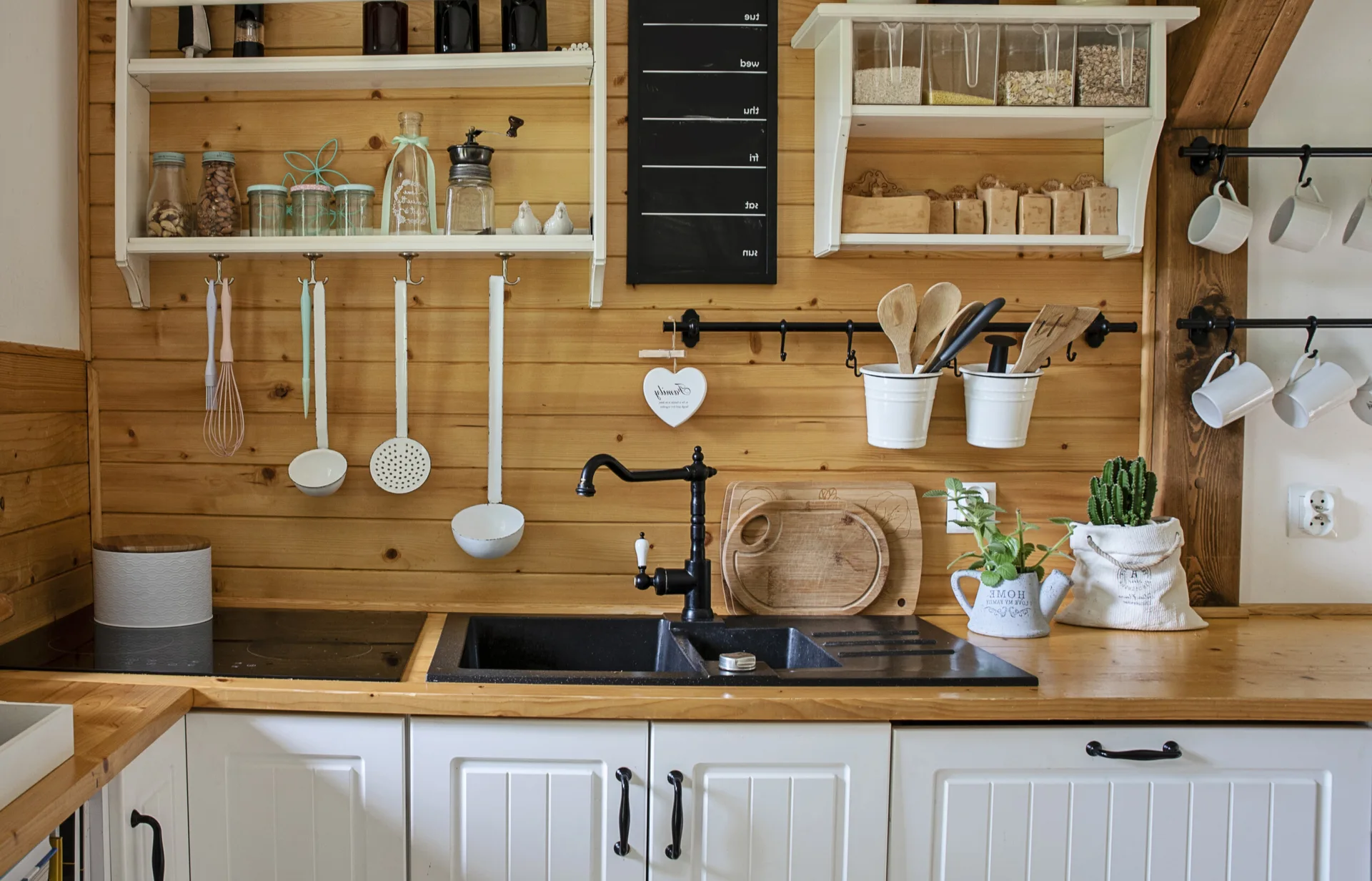 A rustic kitchen counter features a black double sink, wooden countertops, white shaker cabinets, and horizontal wood-paneled walls with open shelving.