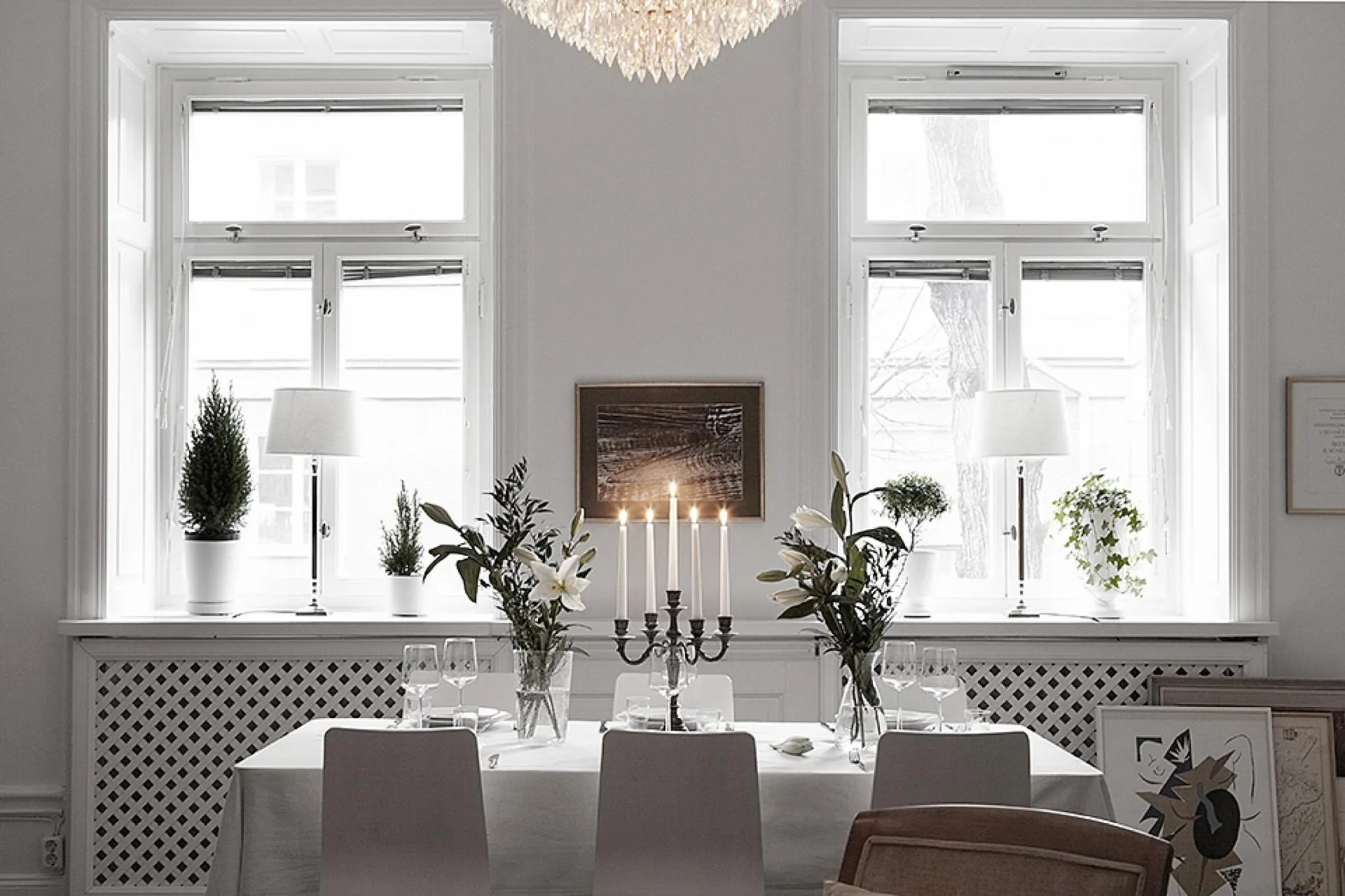 A white dining room features a set table with a candelabra in front of two large windows with decorative lattice radiator covers.