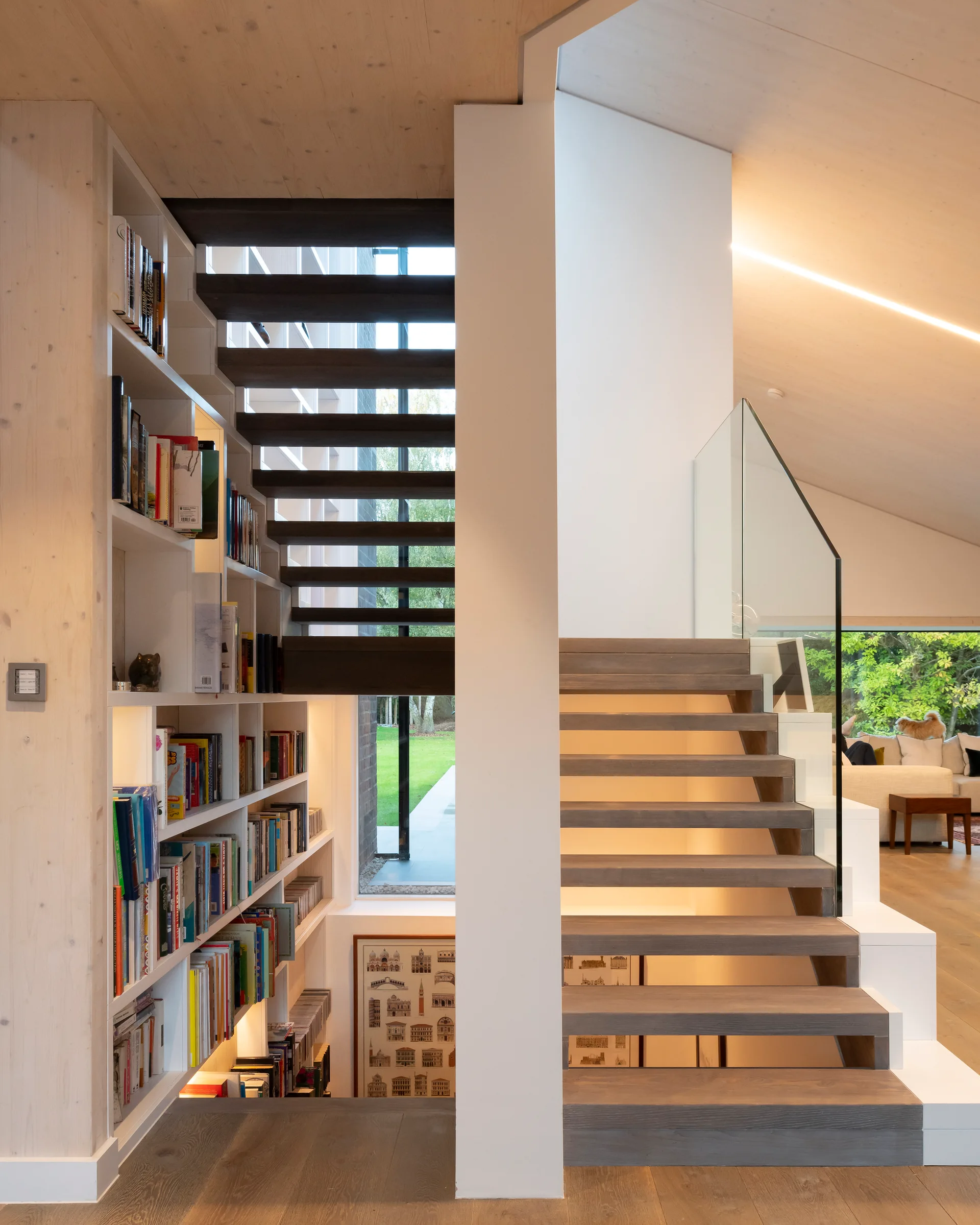 A minimalist wooden staircase with open treads and a glass railing is integrated with tall built-in bookshelves lining the white stairwell walls.