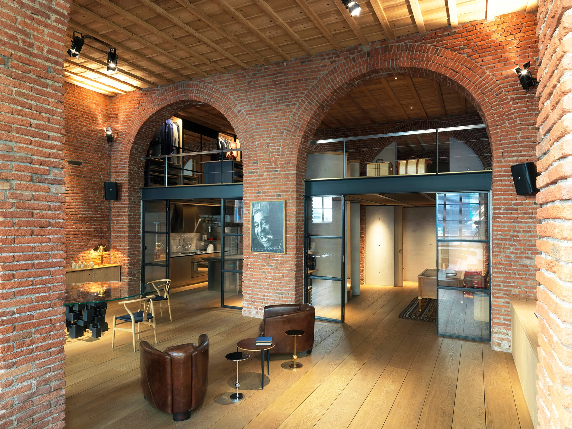An industrial loft living space featuring large red brick arches, wide-plank wood floors, exposed wood ceilings, and black-framed glass partitions.