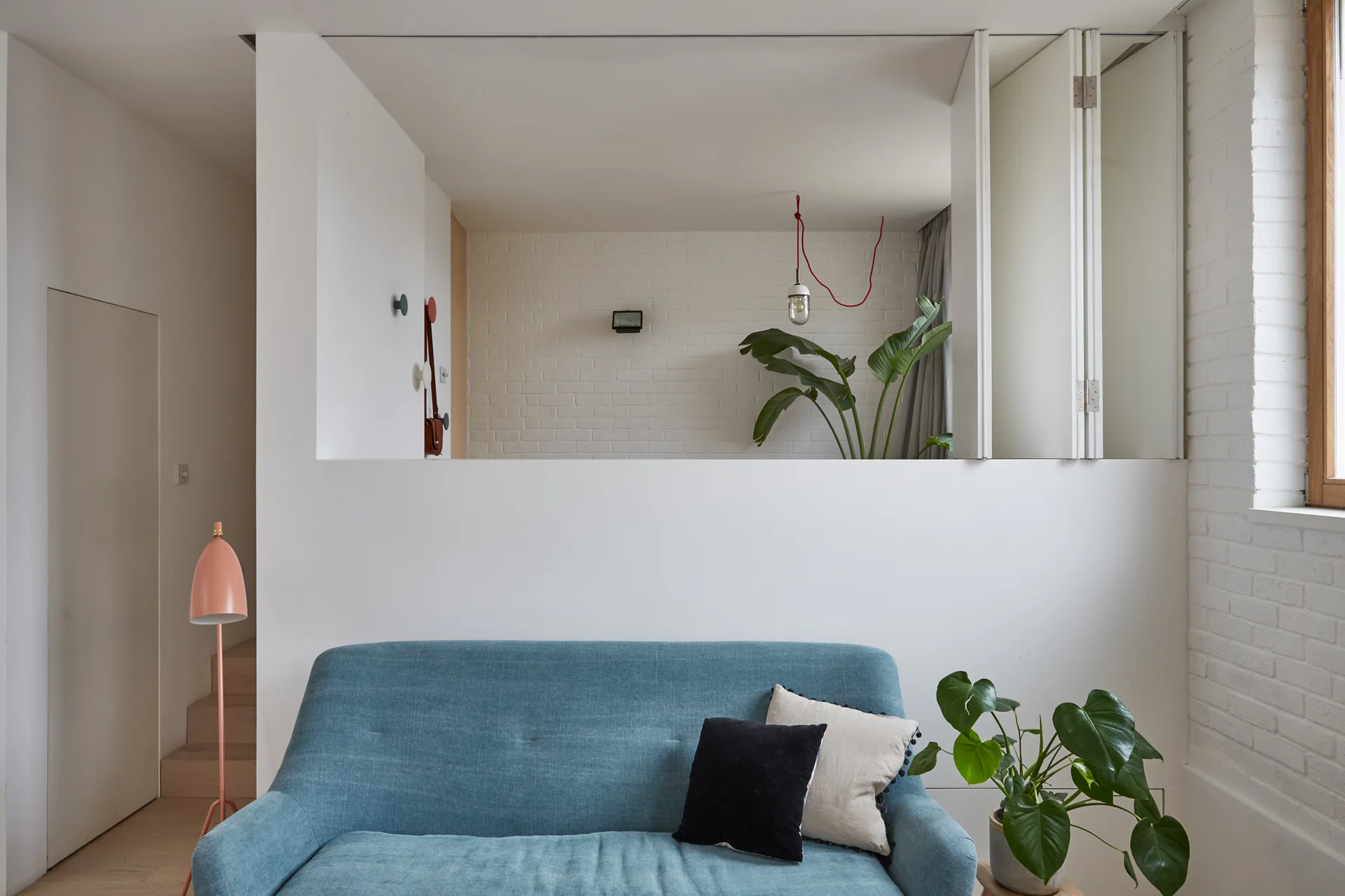 A blue sofa sits against a white interior partition wall with a large cutout opening that looks into an adjacent room with painted brick.