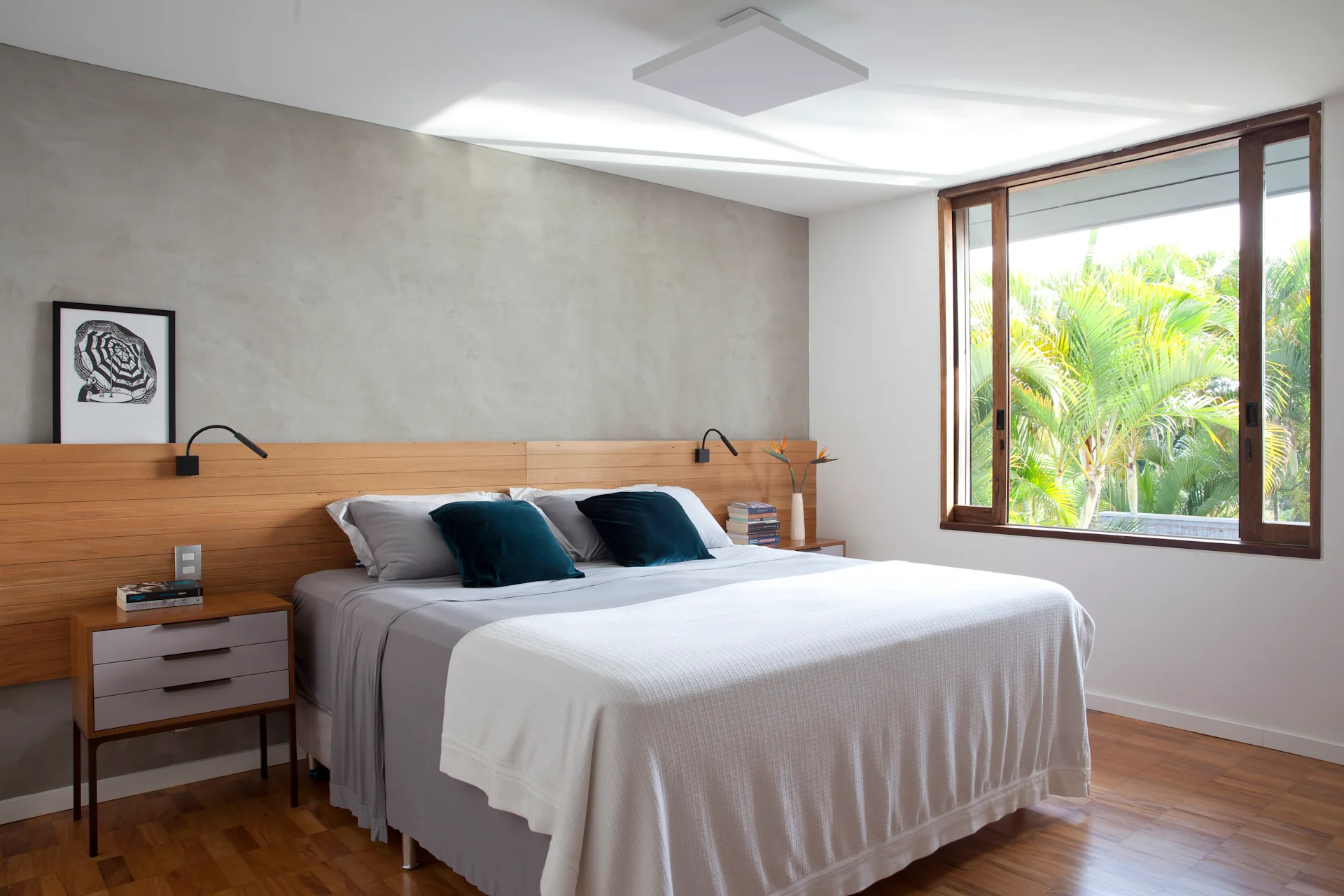 A modern bedroom features a low wood paneled headboard wall against a gray textured concrete wall, a large window, and gray bedding.