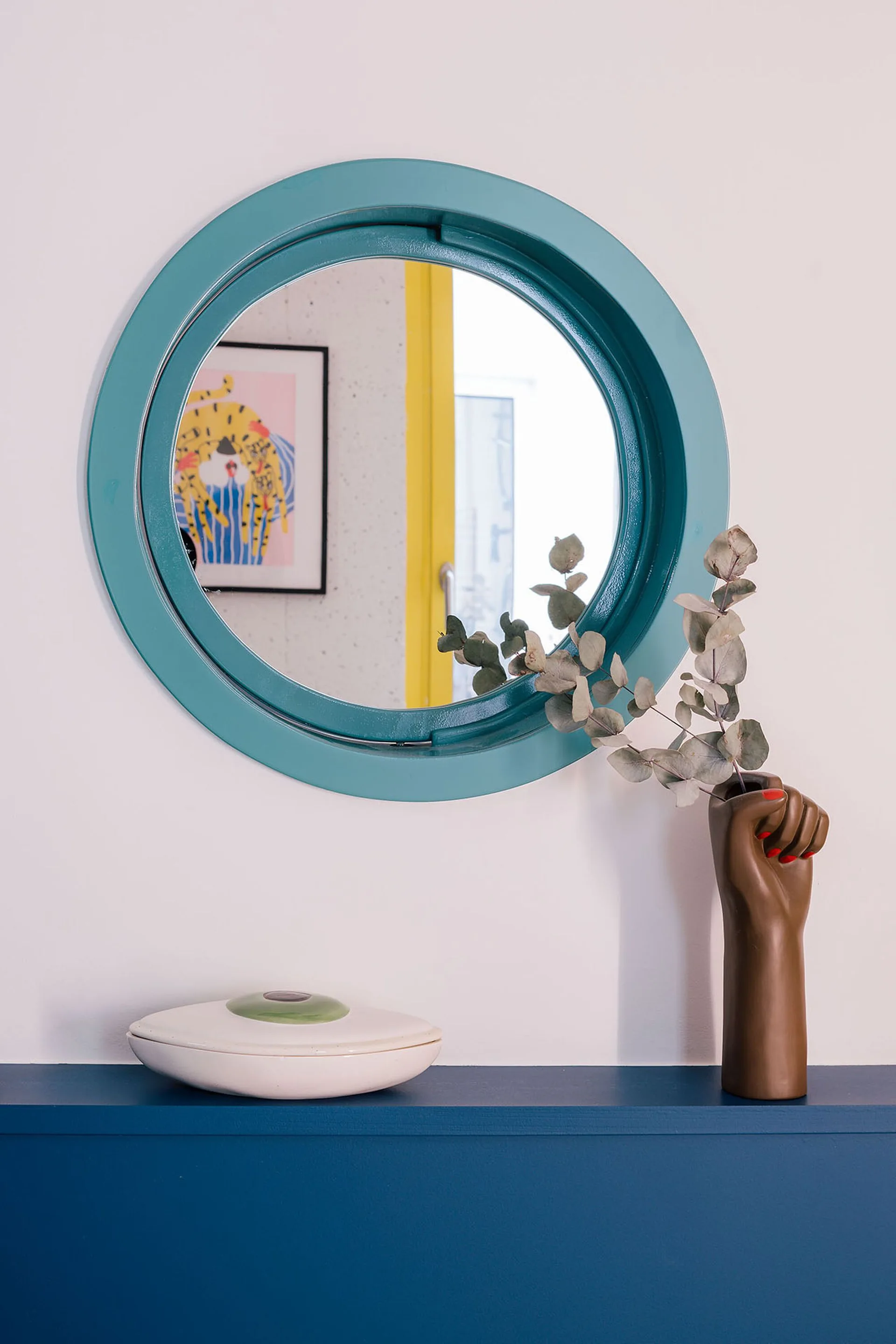 A round teal-framed mirror hangs above a dark blue shelf styled with a hand-shaped brown ceramic vase holding eucalyptus and a white lidded box.