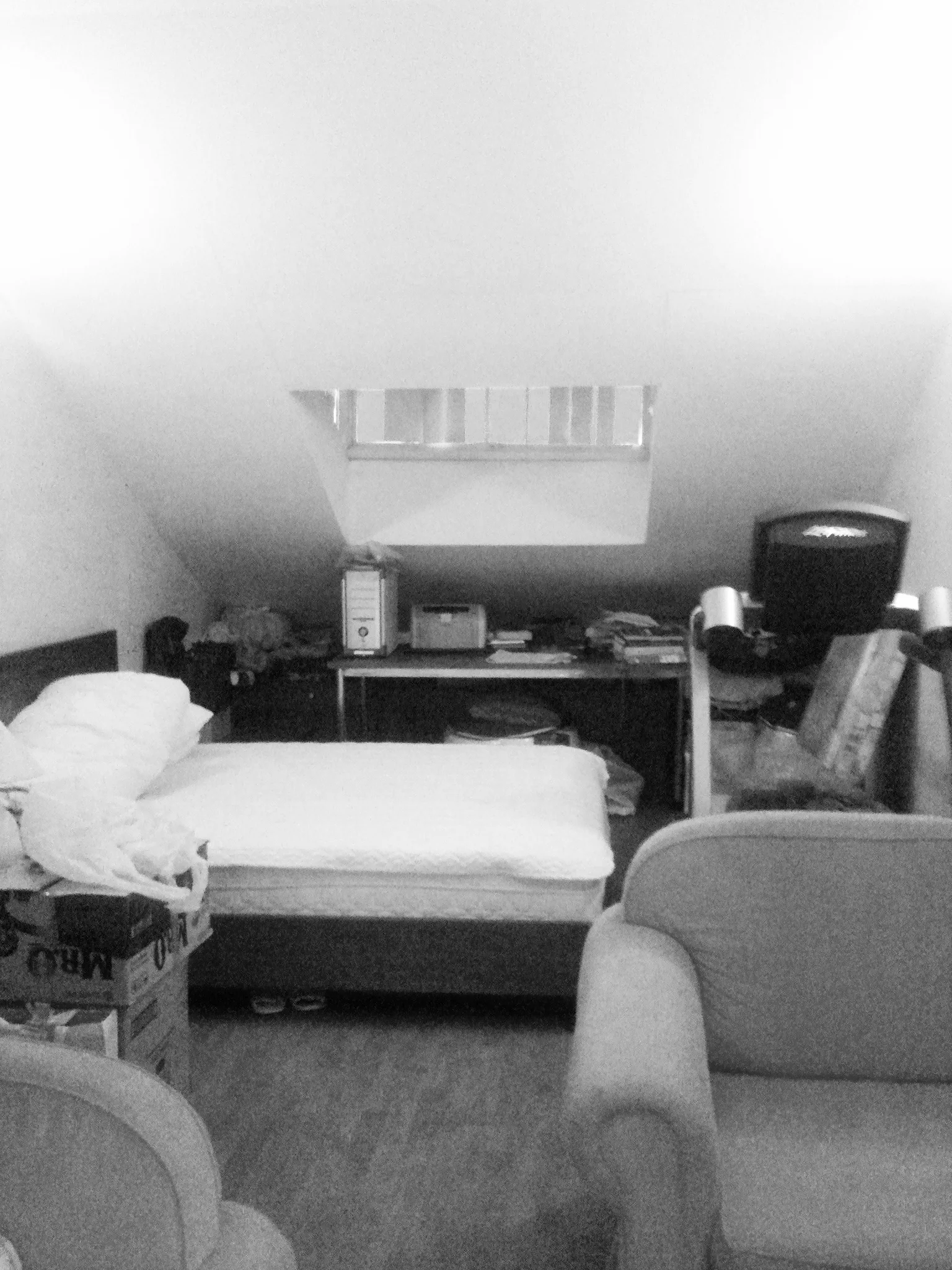 A black and white photo of an attic bedroom with a low bed, a desk placed under a skylight, and an armchair.