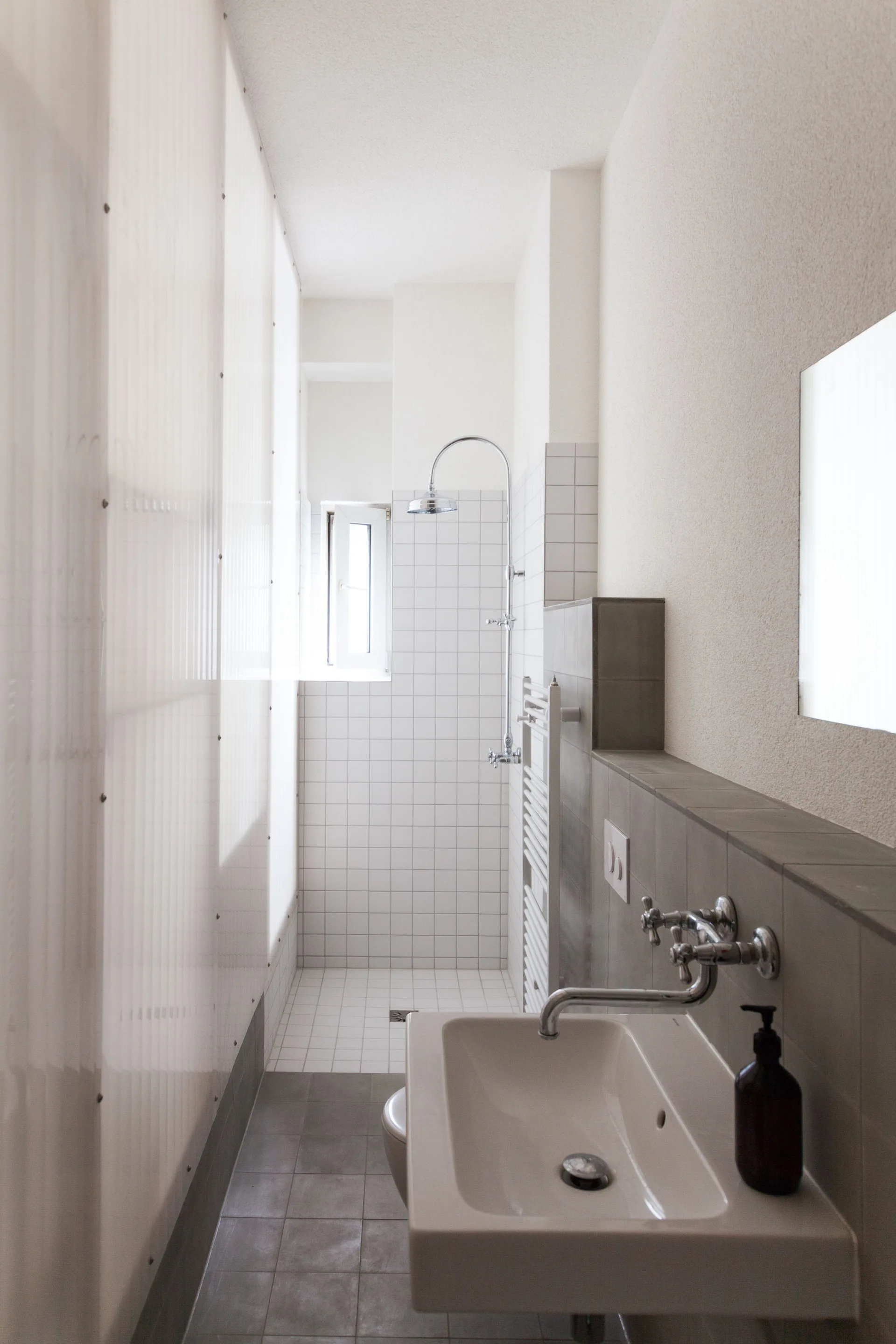 A narrow bathroom featuring a wall-mounted sink with chrome faucet, a white grid-tiled shower, and a translucent corrugated partition wall.