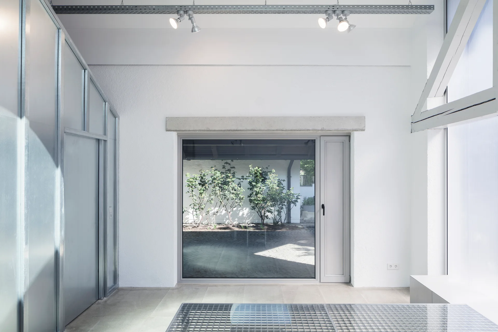 A minimalist industrial room with white textured plaster walls, a large glass patio door, industrial track lighting, and metallic wall panels.