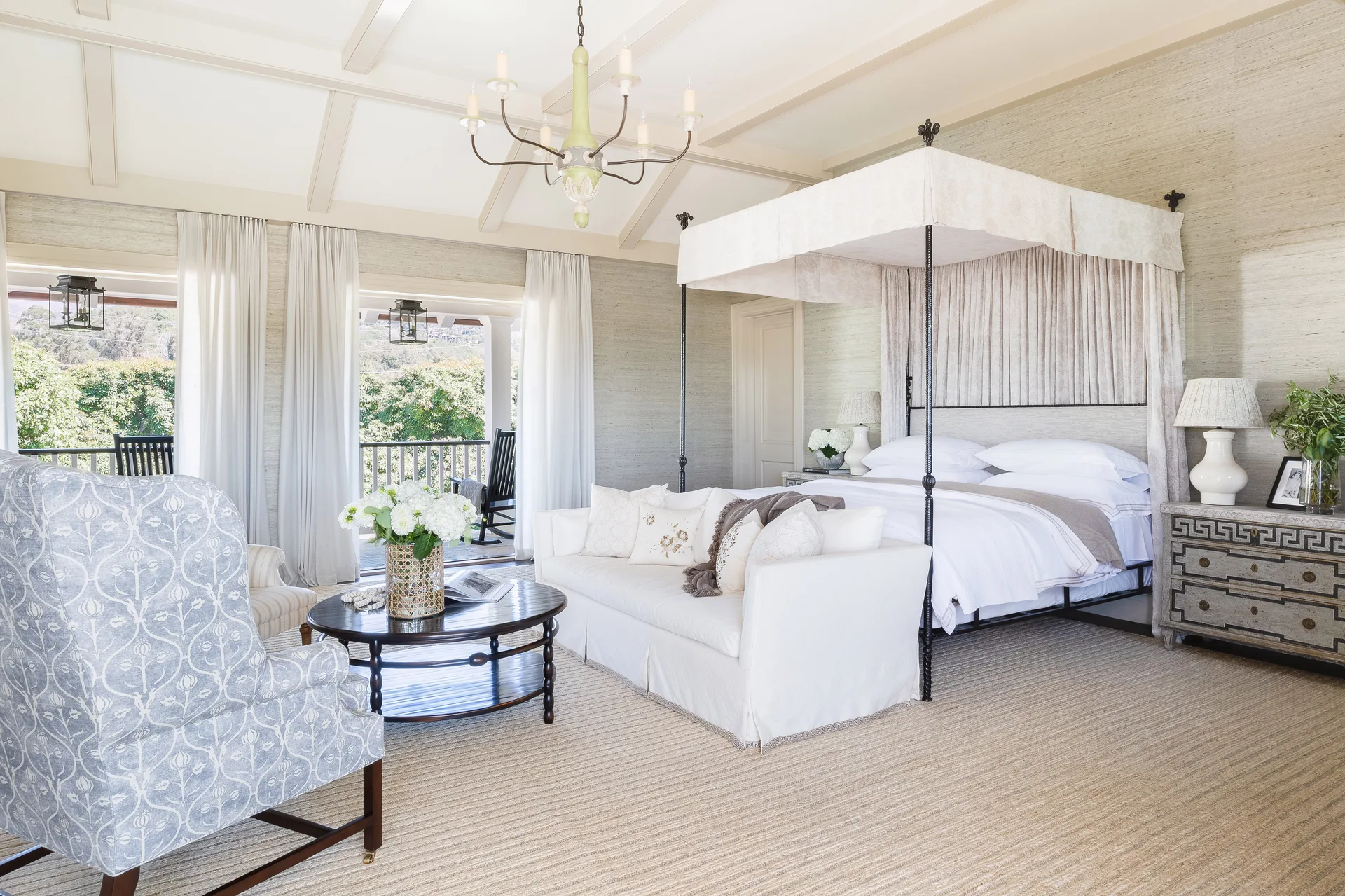 A spacious bedroom featuring a black iron canopy bed, a white sofa at its foot, a patterned armchair, and vaulted beamed ceilings.