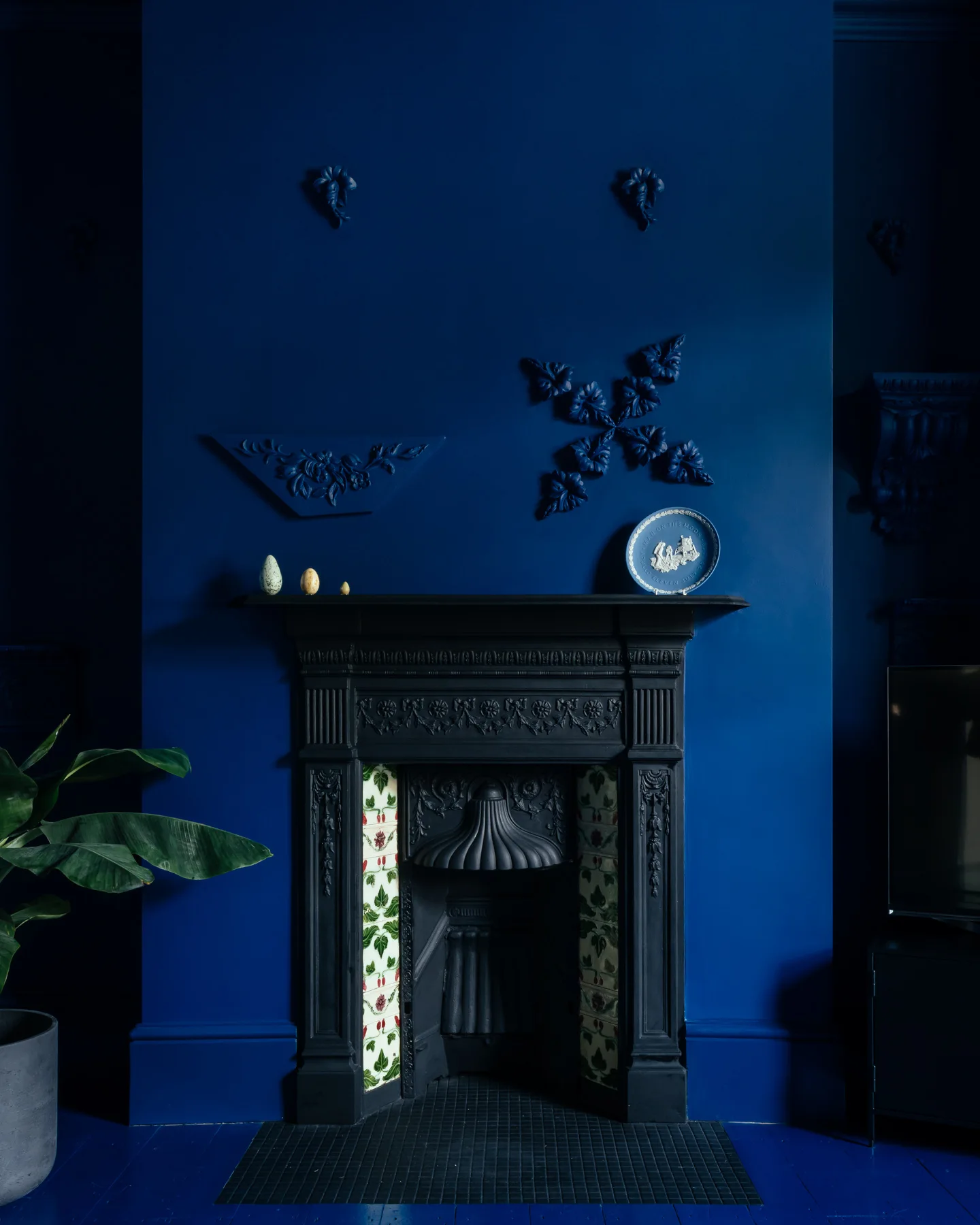 An ornate black cast iron fireplace with floral tile inserts is set against a deep blue wall decorated with matching blue plaster moldings.