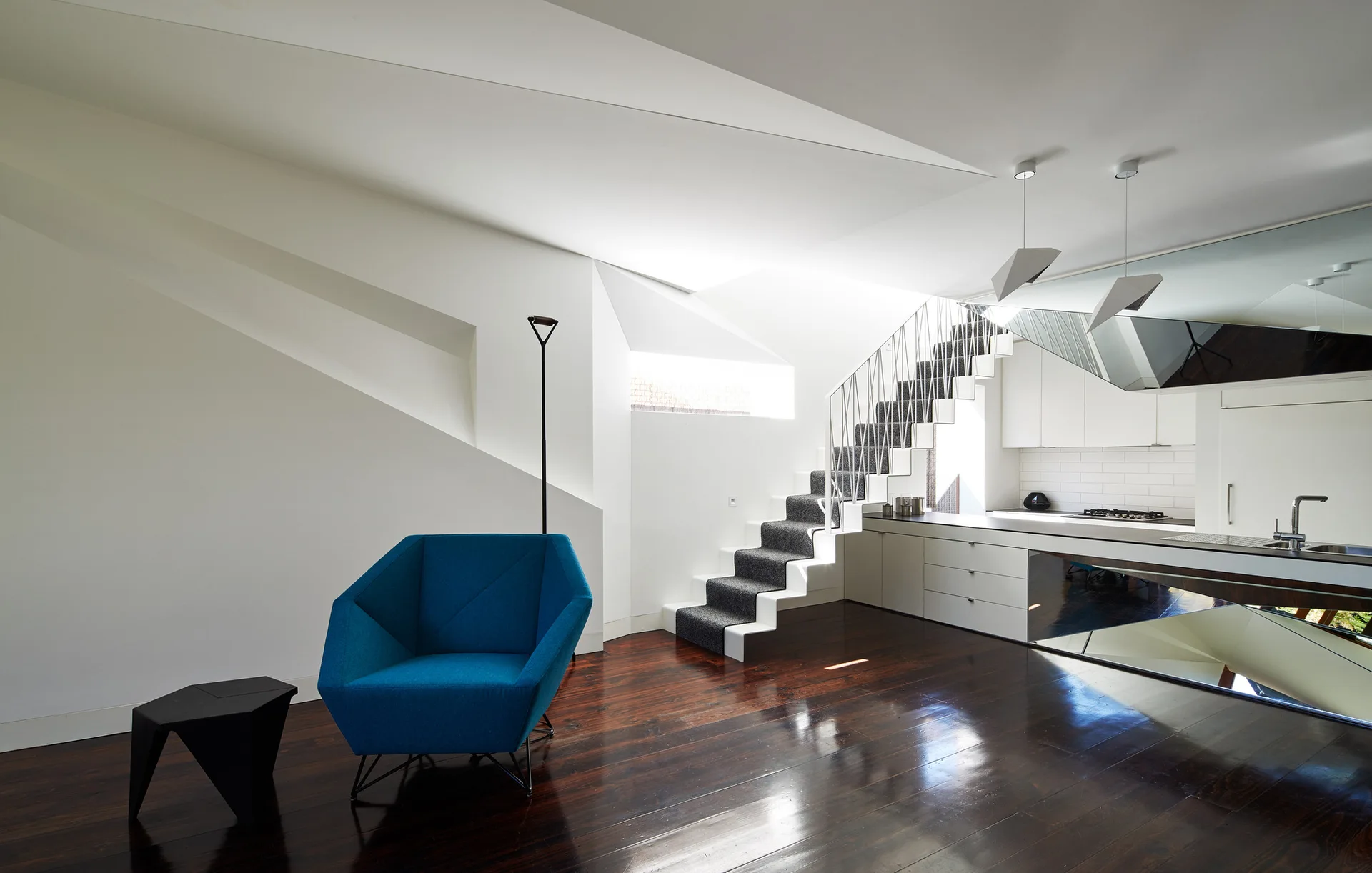 A modern open-plan living room features an angular blue armchair, glossy dark wood floors, and a white geometric staircase.