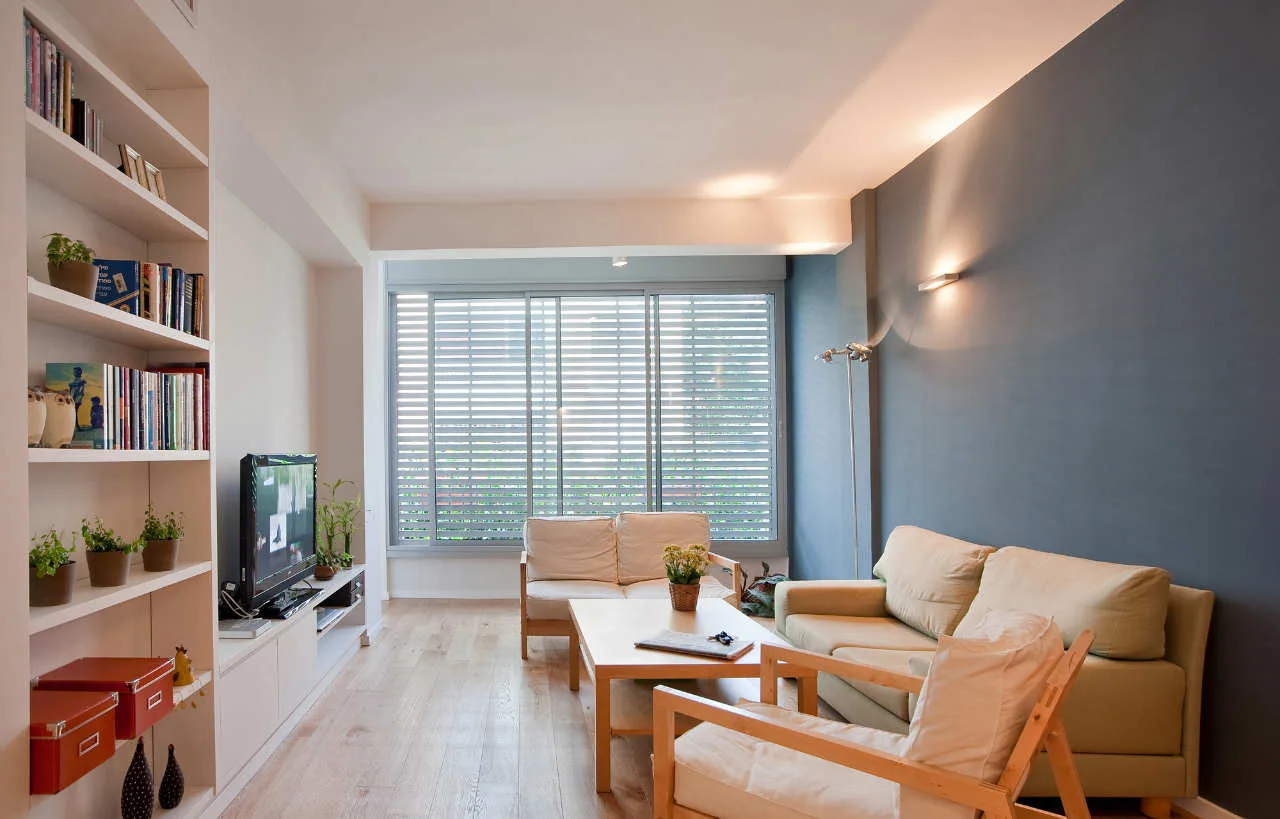 A living room features a dark blue-grey accent wall, light wood flooring, white built-in shelving, and light wood framed sofas.