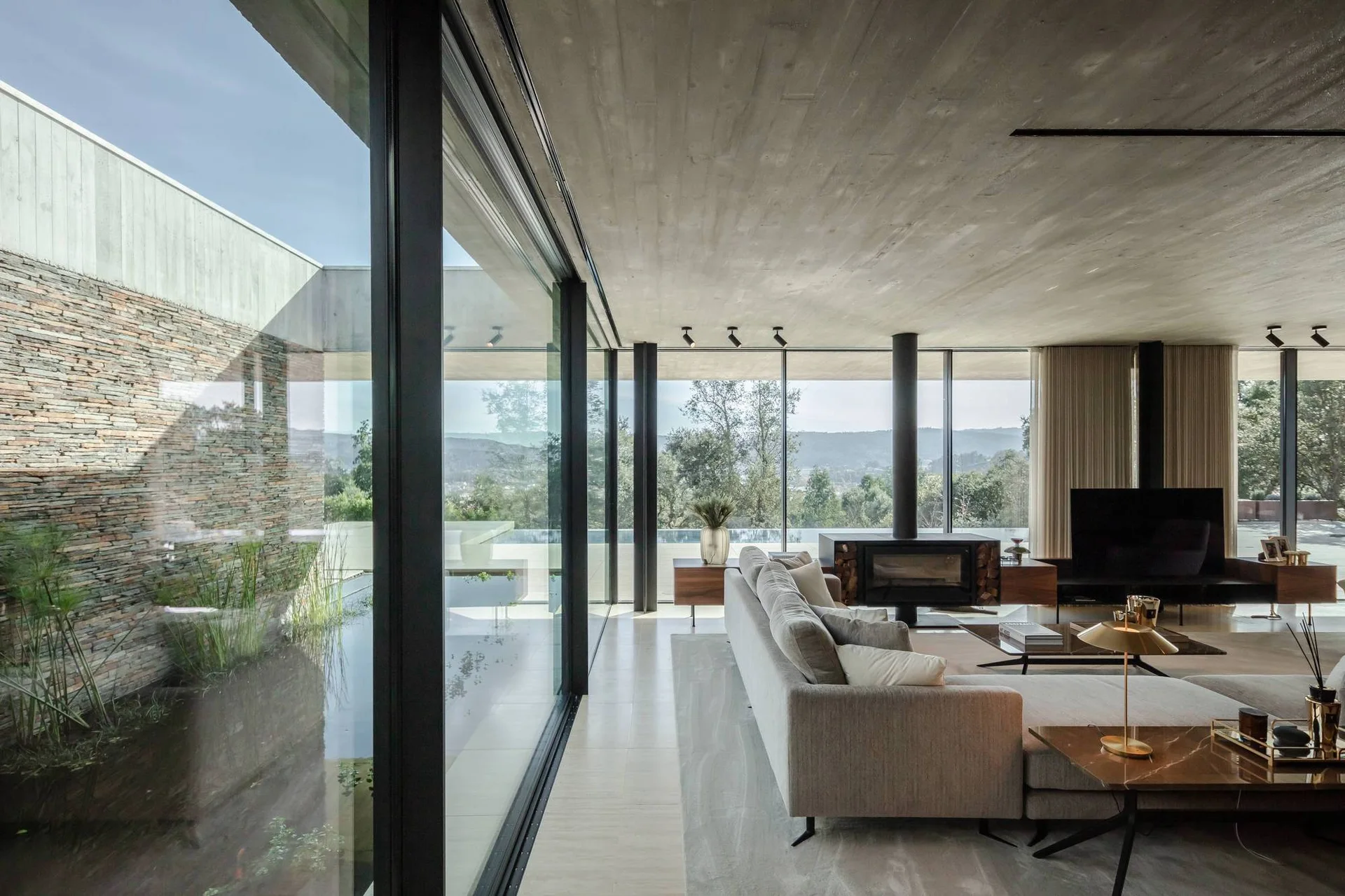 A modern living room features a grey modular sofa, a freestanding fireplace, and an exposed concrete ceiling facing large floor-to-ceiling glass walls.