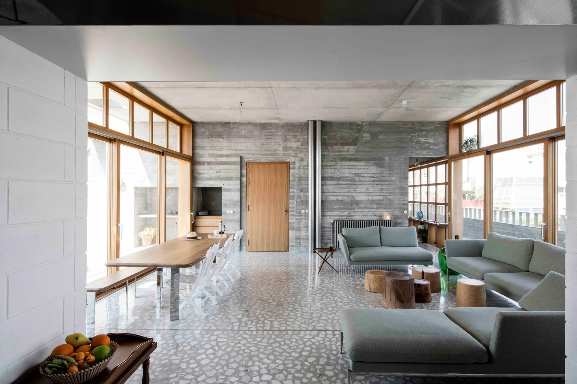A modern open-plan living and dining room with board-formed concrete walls, terrazzo flooring, light green sofas, and a long wooden dining table.