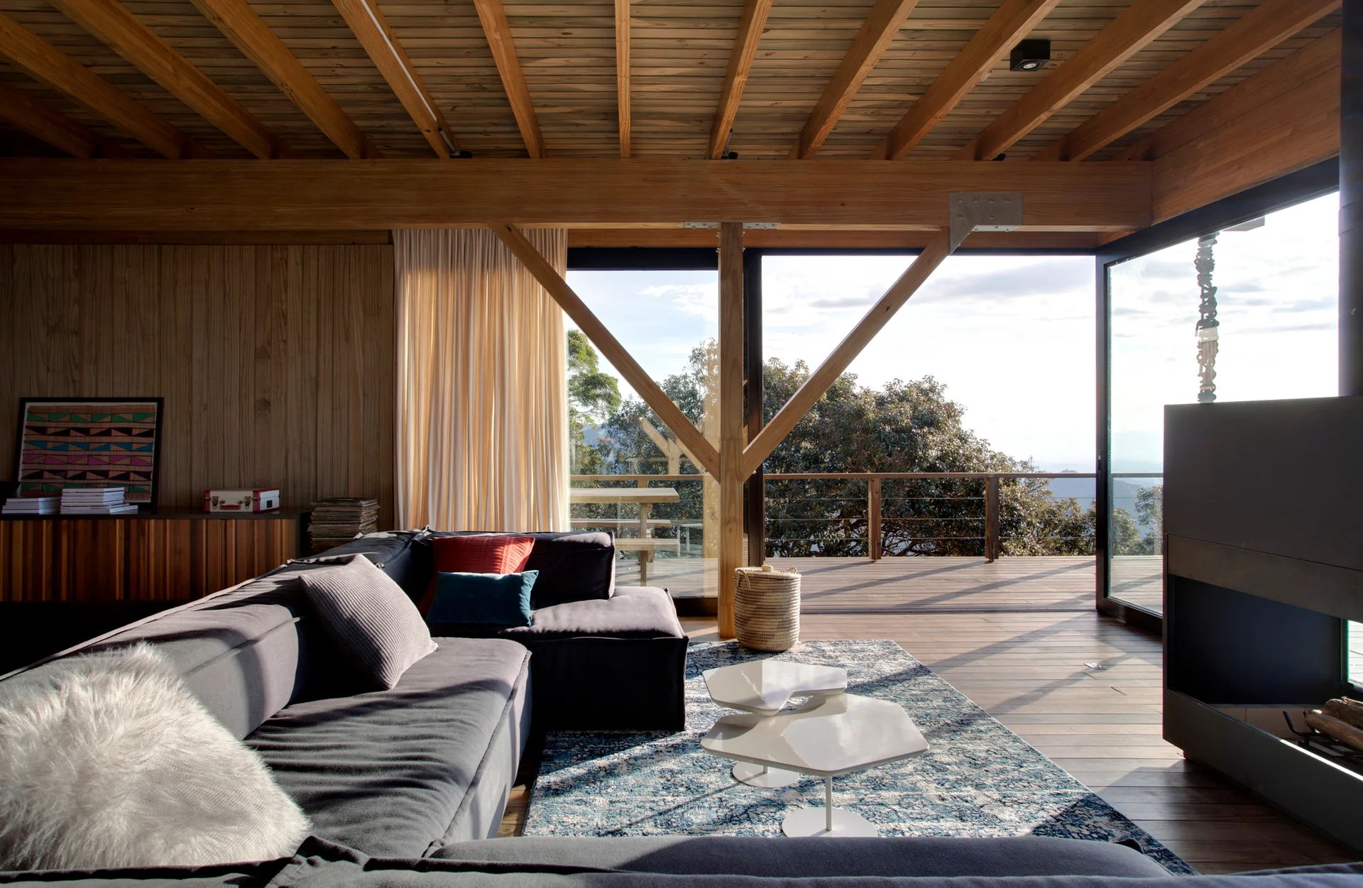 A rustic contemporary living room features a grey sectional, a blue patterned rug, exposed timber ceiling beams, and sliding doors opening to a wood deck.
