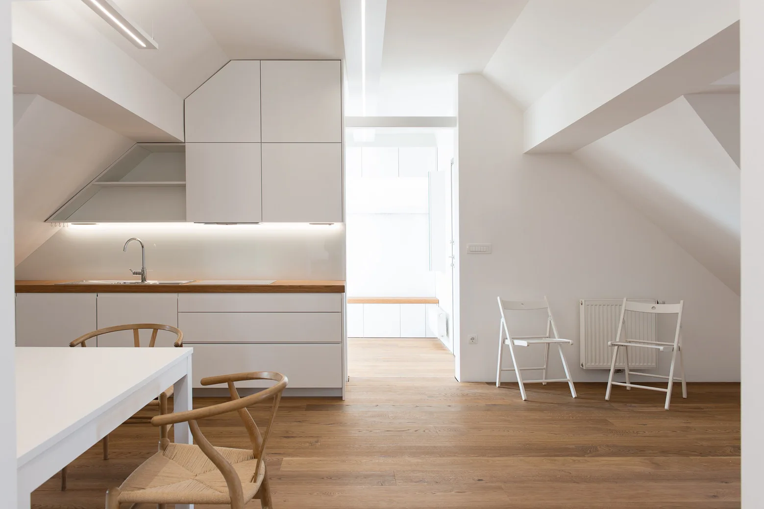 A minimalist attic kitchen with custom white cabinetry cut to fit a sloped ceiling, wood countertops, and light wood floors.