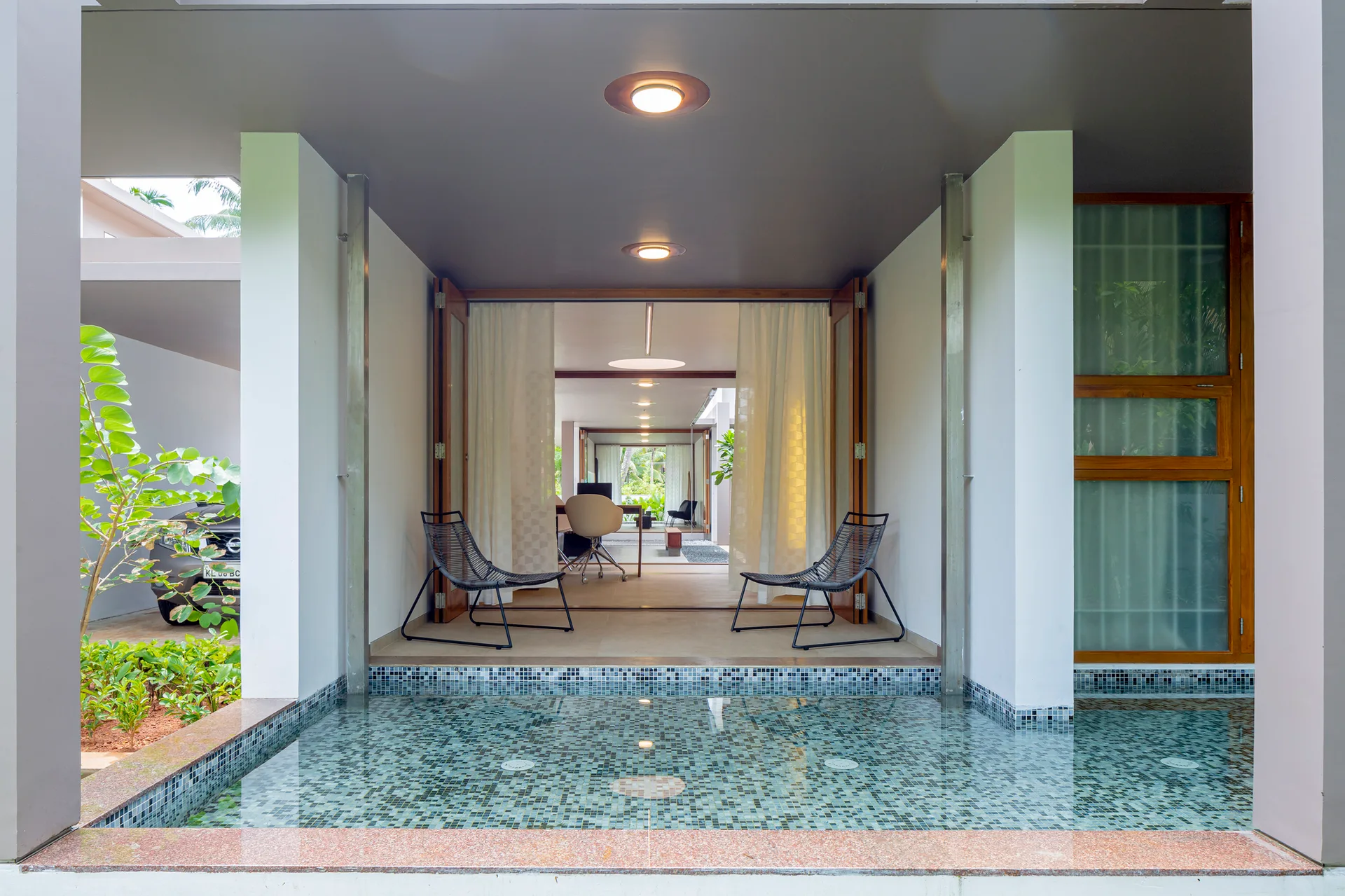A covered patio featuring a shallow mosaic-tiled pool, two black wire-frame lounge chairs, and wooden folding doors opening to the interior.