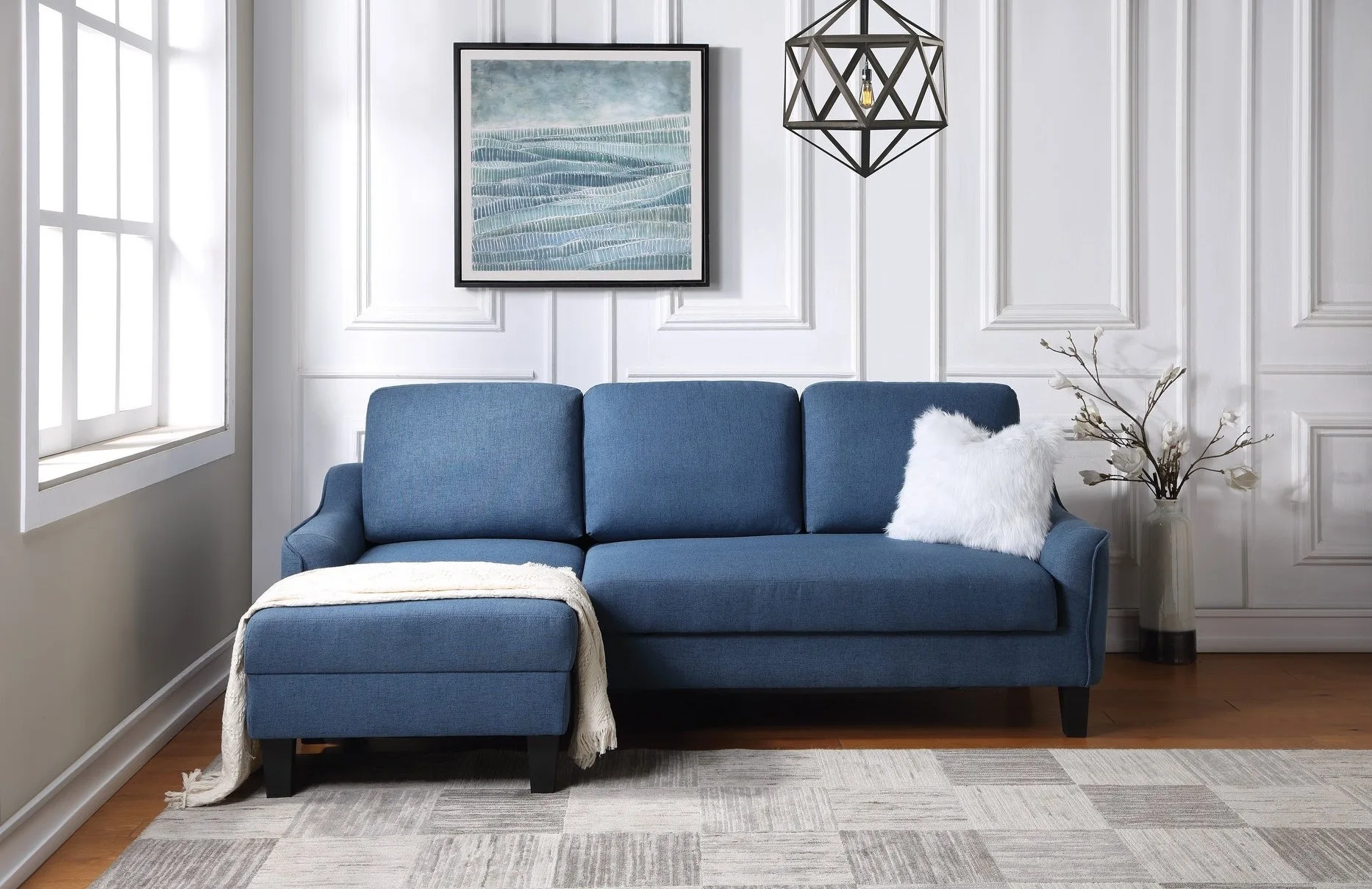A blue fabric sofa with a matching ottoman sits on a patterned gray rug against a white paneled wall decorated with a framed painting.