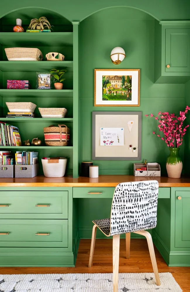 A home office features built-in green cabinetry and arched shelves above a warm wood desk paired with a patterned chair.