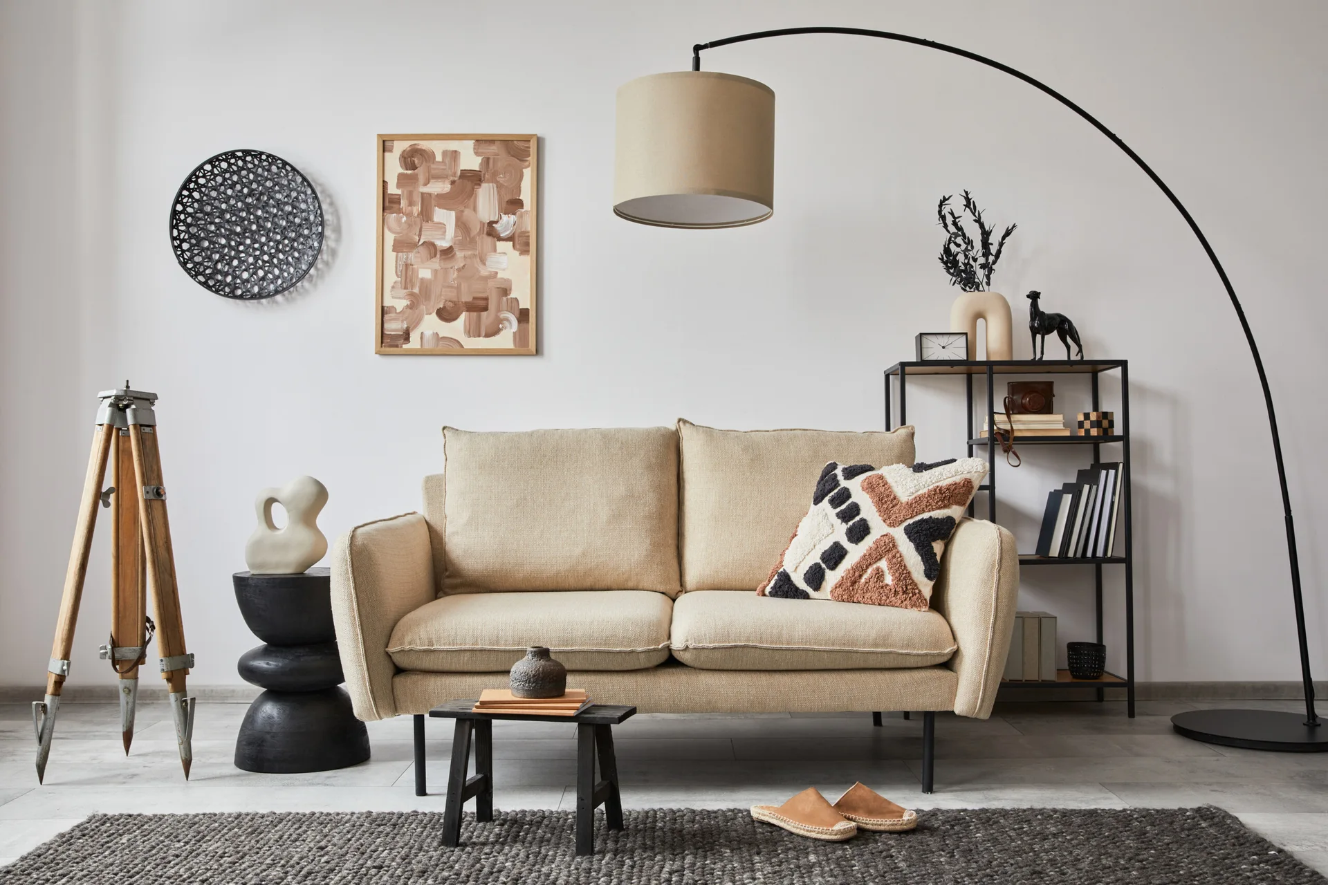 A beige fabric sofa sits on a textured grey rug, flanked by a black arched floor lamp, a metal display shelf, and a vintage wooden tripod.