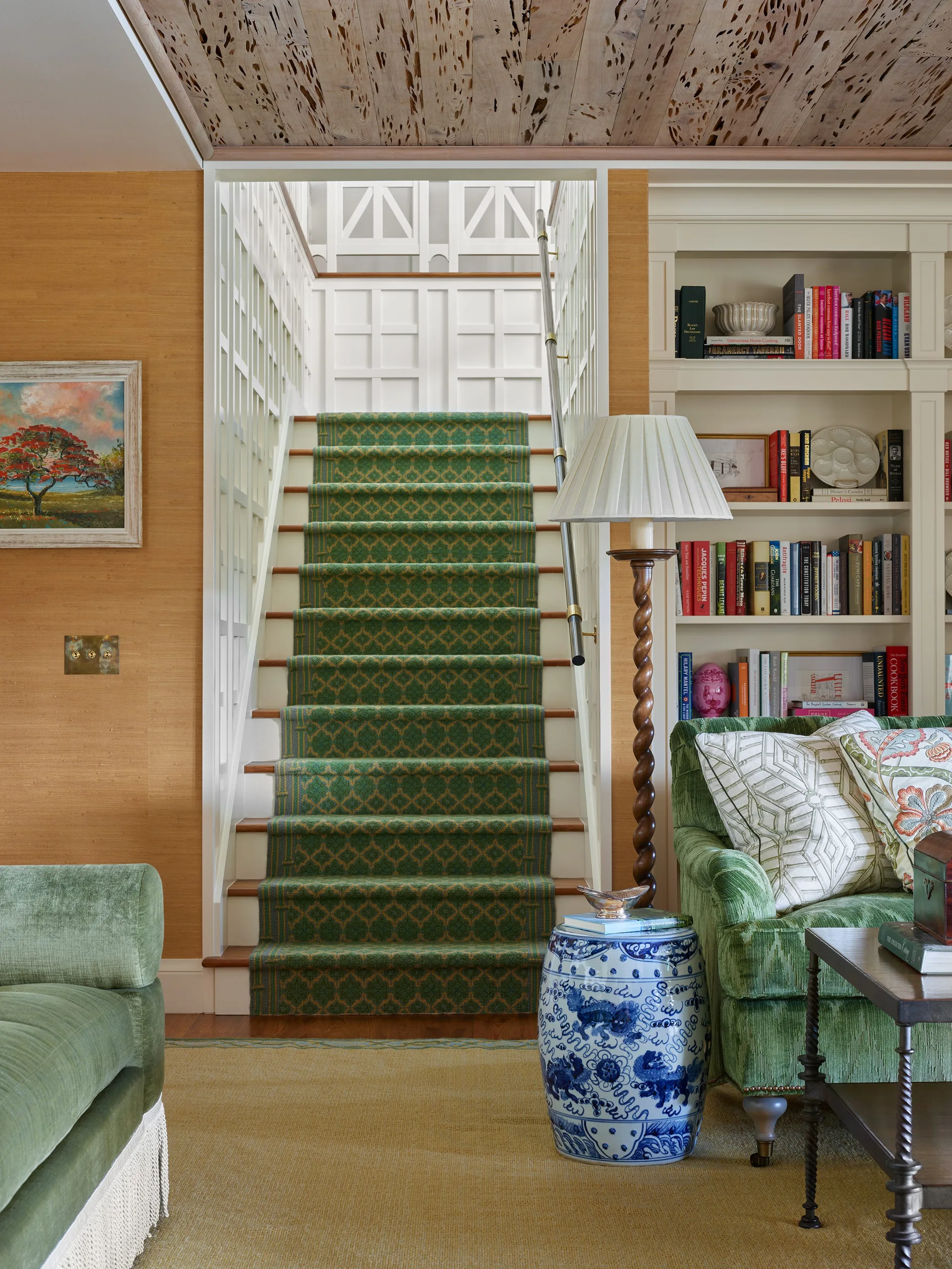 A traditional living room features a staircase with a green patterned runner, built-in bookshelves, and a green velvet sofa.