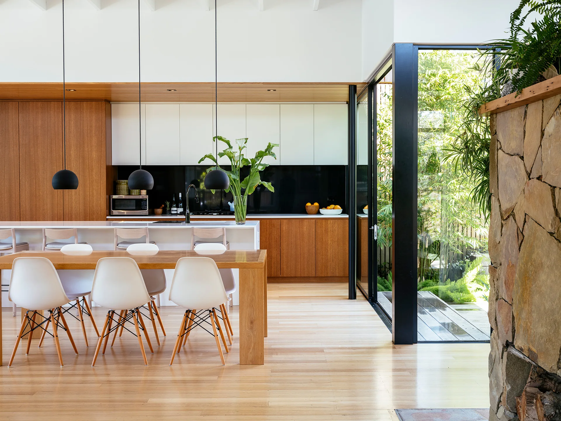 A modern open-concept kitchen and dining area with wood cabinetry, a wooden table, white chairs, and a glass wall opening to a green garden.