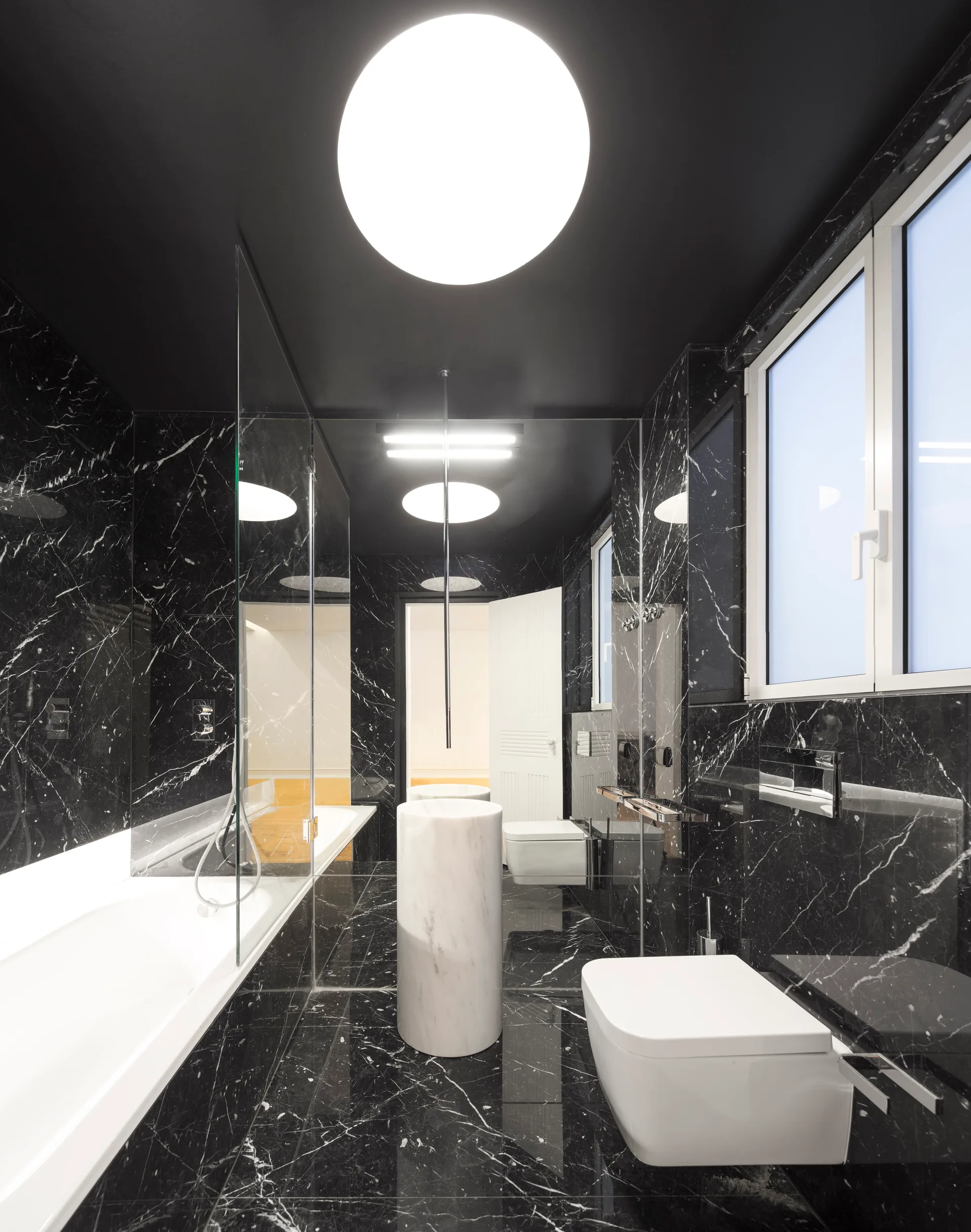 A modern bathroom featuring black veined marble walls and floors, a white cylindrical pedestal sink, and a wall-mounted toilet.