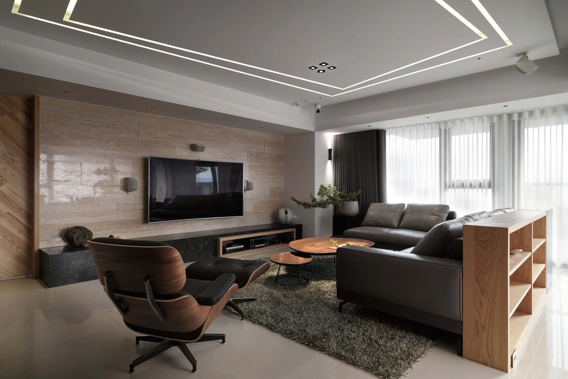 A modern living room features a travertine stone accent wall with a mounted TV, a grey leather sofa, and a classic wood lounge chair.